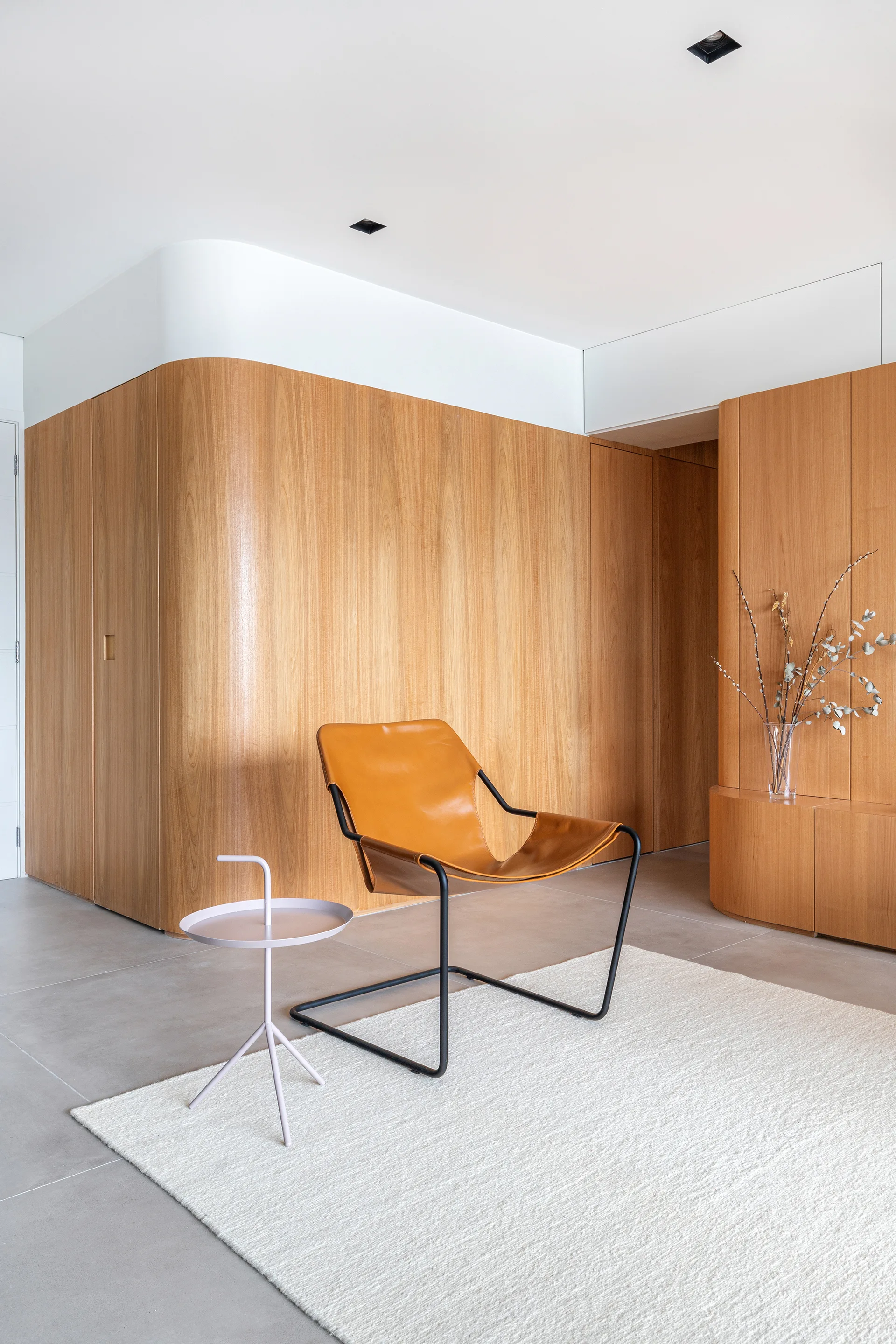 A tan leather sling chair sits on an off-white rug next to a light purple side table, backed by a curved wood-paneled wall.