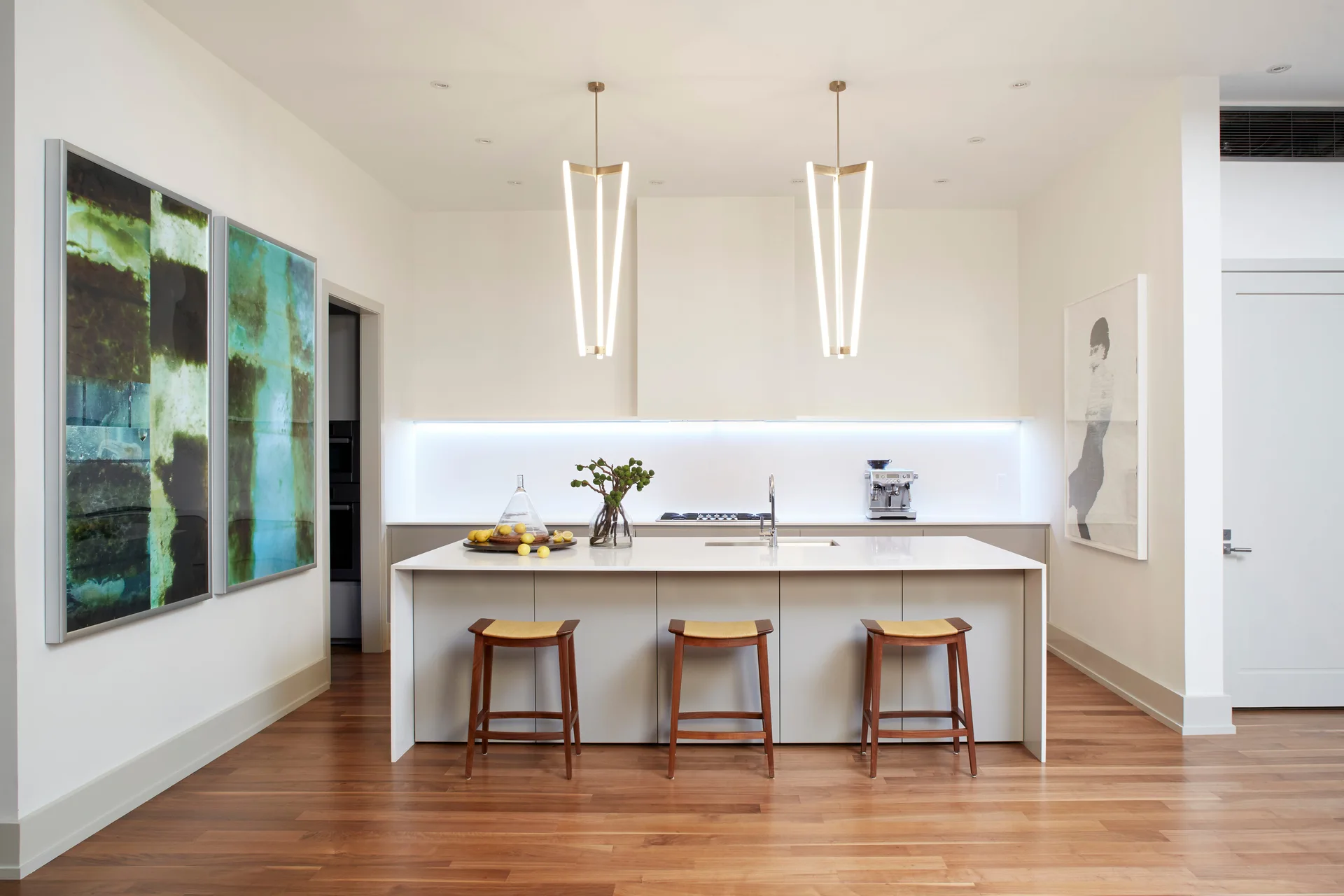 A minimalist kitchen featuring a light gray island with wood stools, vertical tube pendant lights, under-cabinet lighting, and warm hardwood floors.