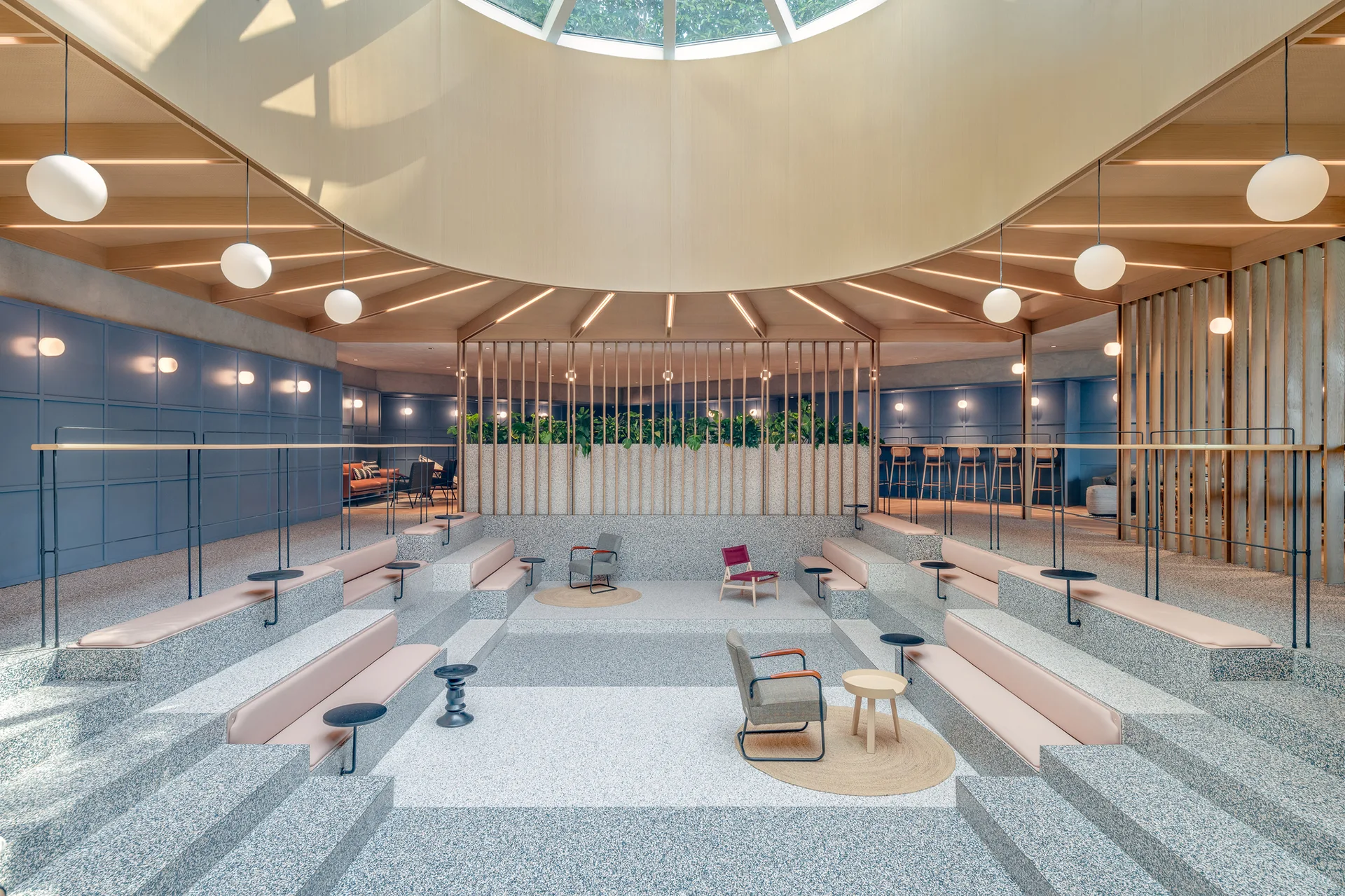 A sunken lounge featuring tiered terrazzo seating with pink cushions, grey armchairs, blue paneled walls, and a large circular skylight overhead.