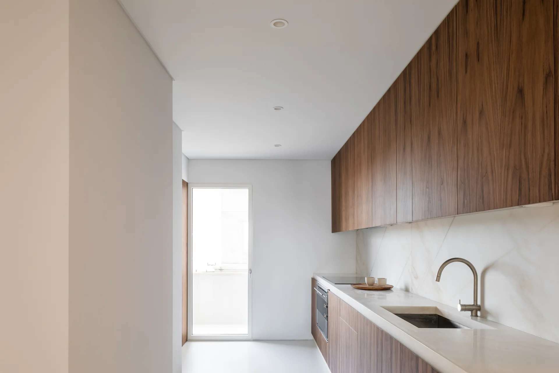 A minimalist galley kitchen features flat-panel walnut cabinetry, a veined stone backsplash and countertop, and a glass door leading outside.