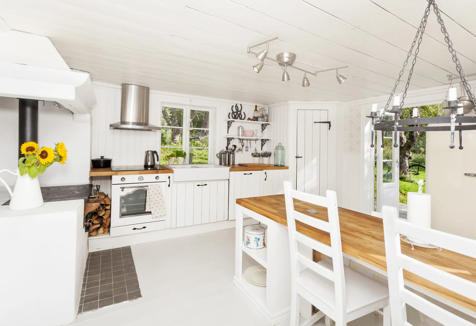 A bright white cottage kitchen features vertical wood paneling, a farmhouse sink, butcher block countertops, and a central island with dining chairs.
