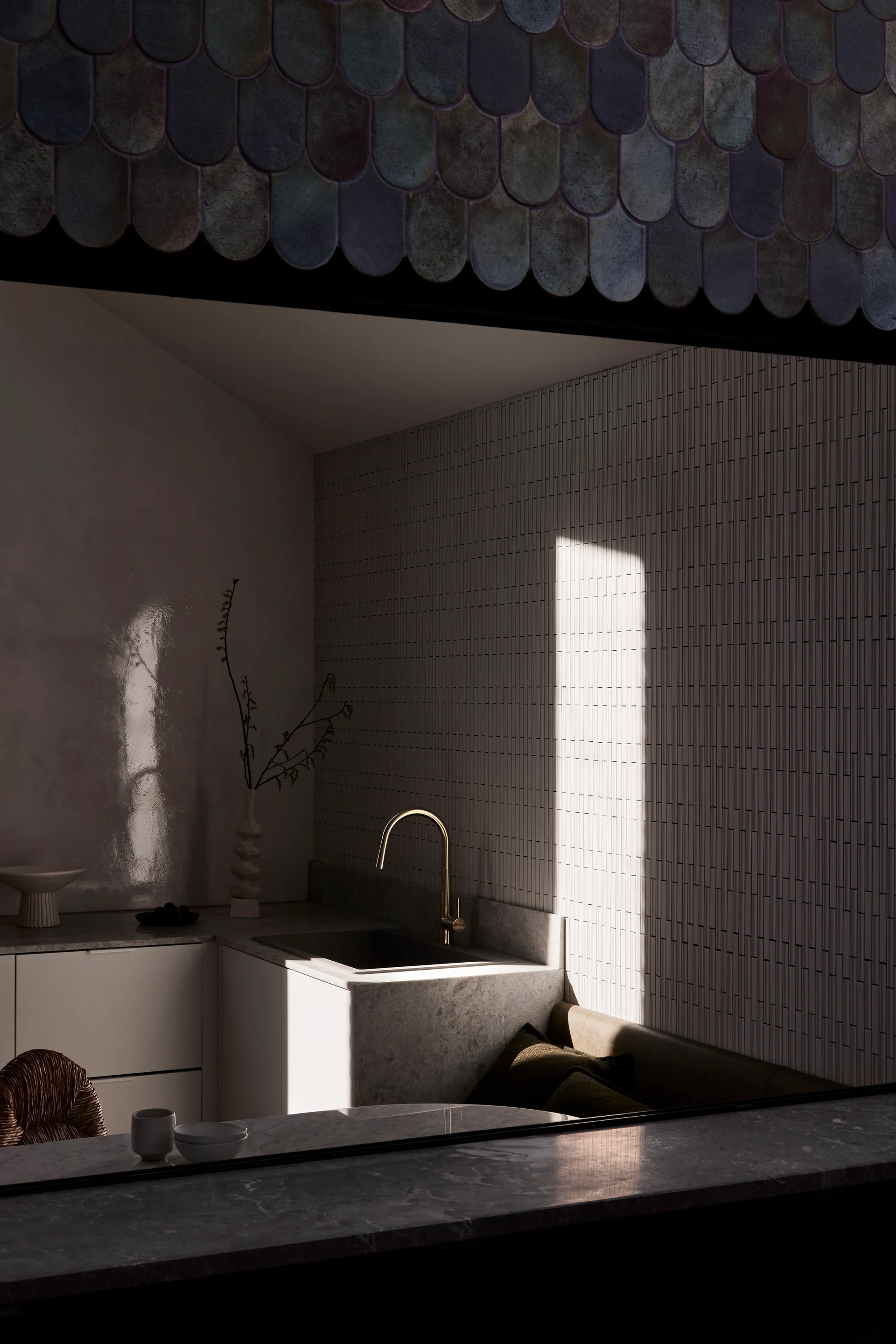A minimalist kitchen features a stone-clad sink with a gold faucet against a vertical white tiled wall.