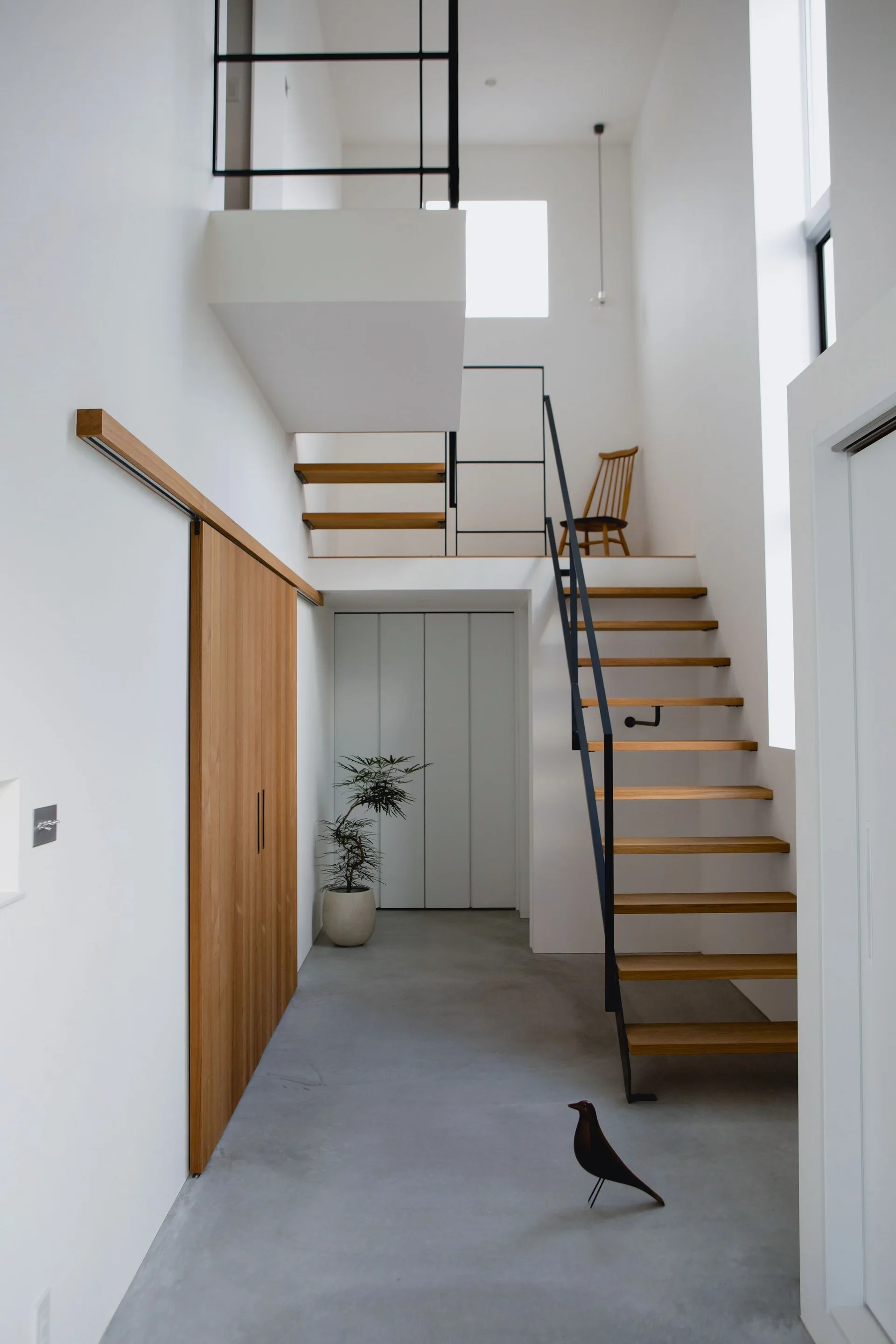 A minimalist entryway featuring a floating wooden staircase, a black metal railing, a sliding wood door, and grey concrete floors.