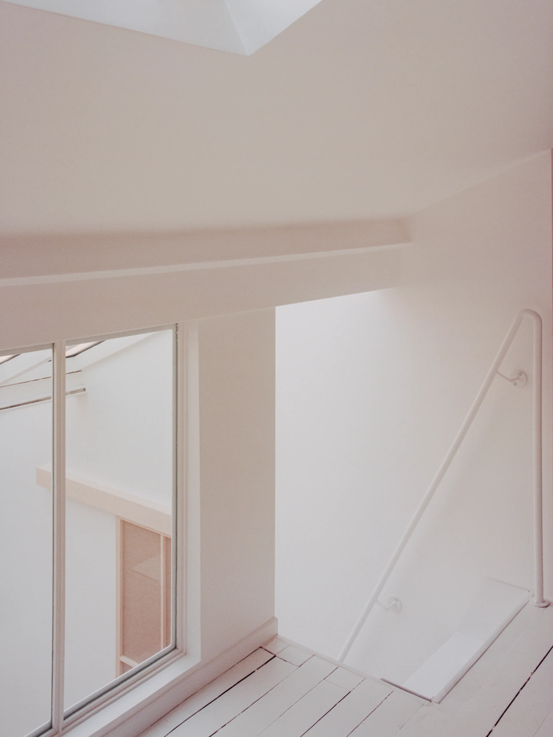 A minimalist white interior space featuring painted wooden floors, a simple white handrail, and an interior window looking into another room.