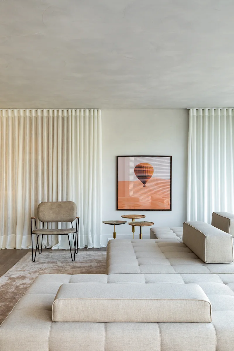 A minimalist living room features a low-profile tufted beige sectional sofa, a modern armchair, and framed artwork depicting a hot air balloon.