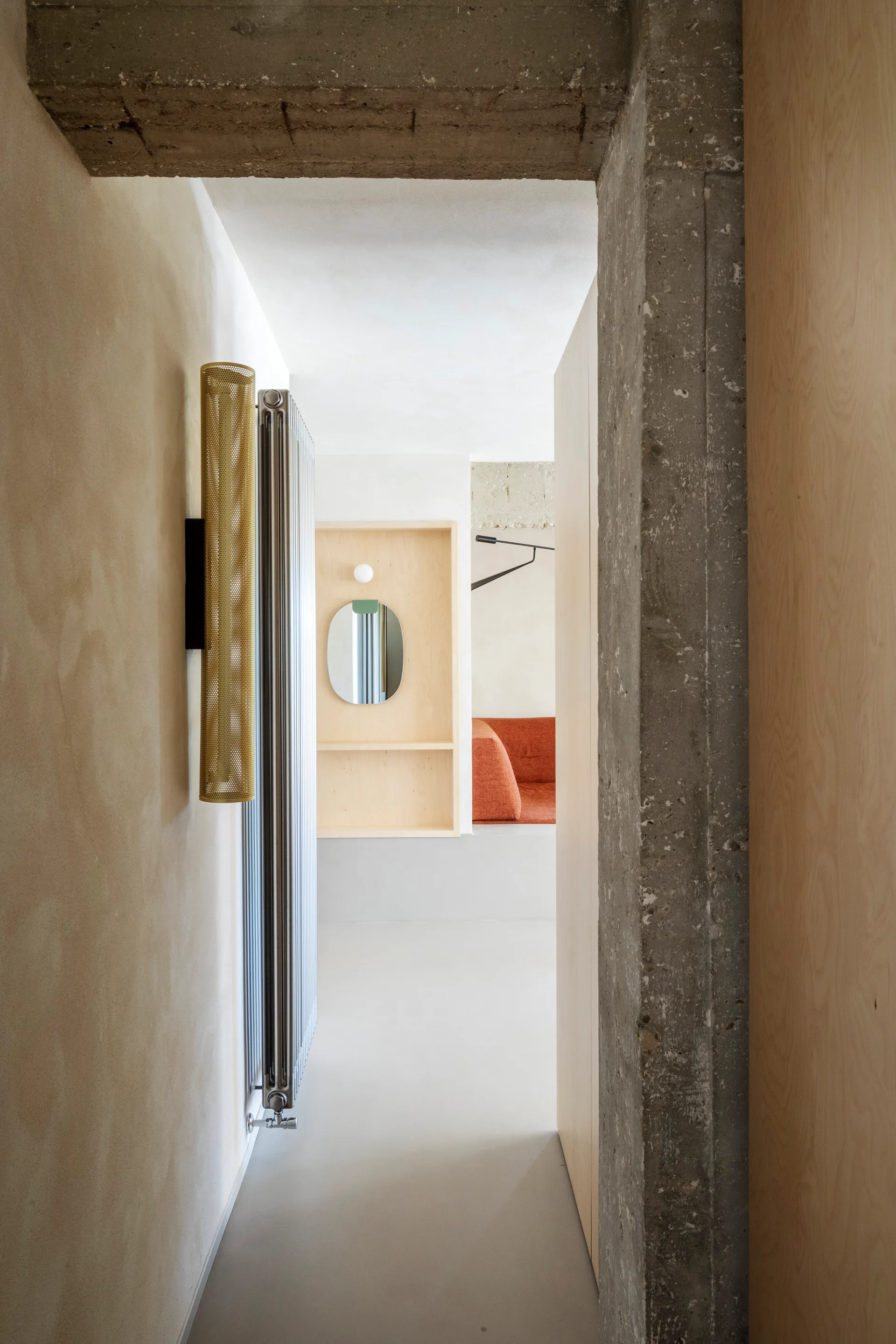 A view down a minimalist hallway with an exposed concrete beam, a vertical metallic radiator, and a light wood wall niche with a small mirror.