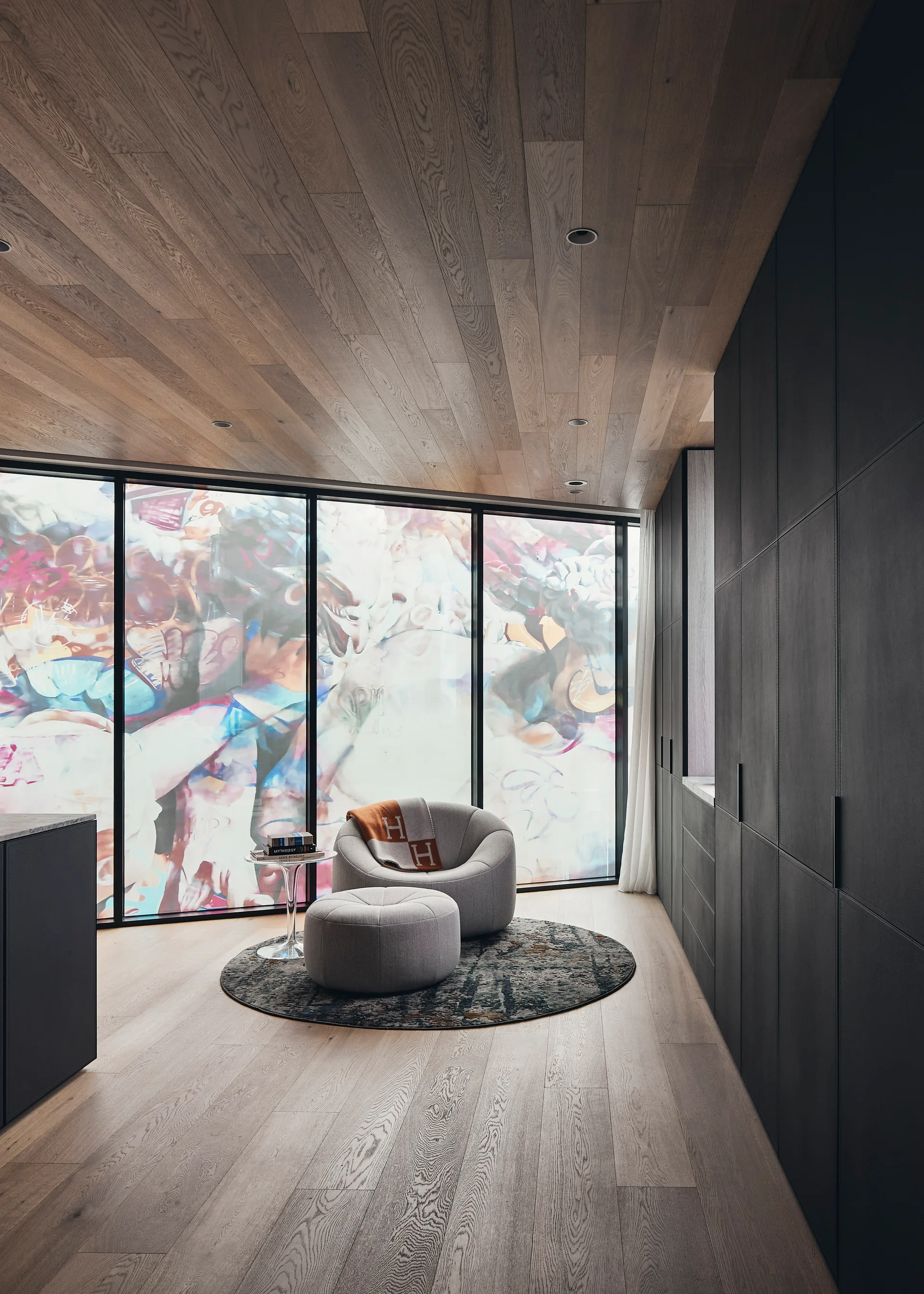 A modern lounge area with a gray fabric armchair and matching ottoman on a round rug with dark built-in cabinets and a wood plank ceiling.