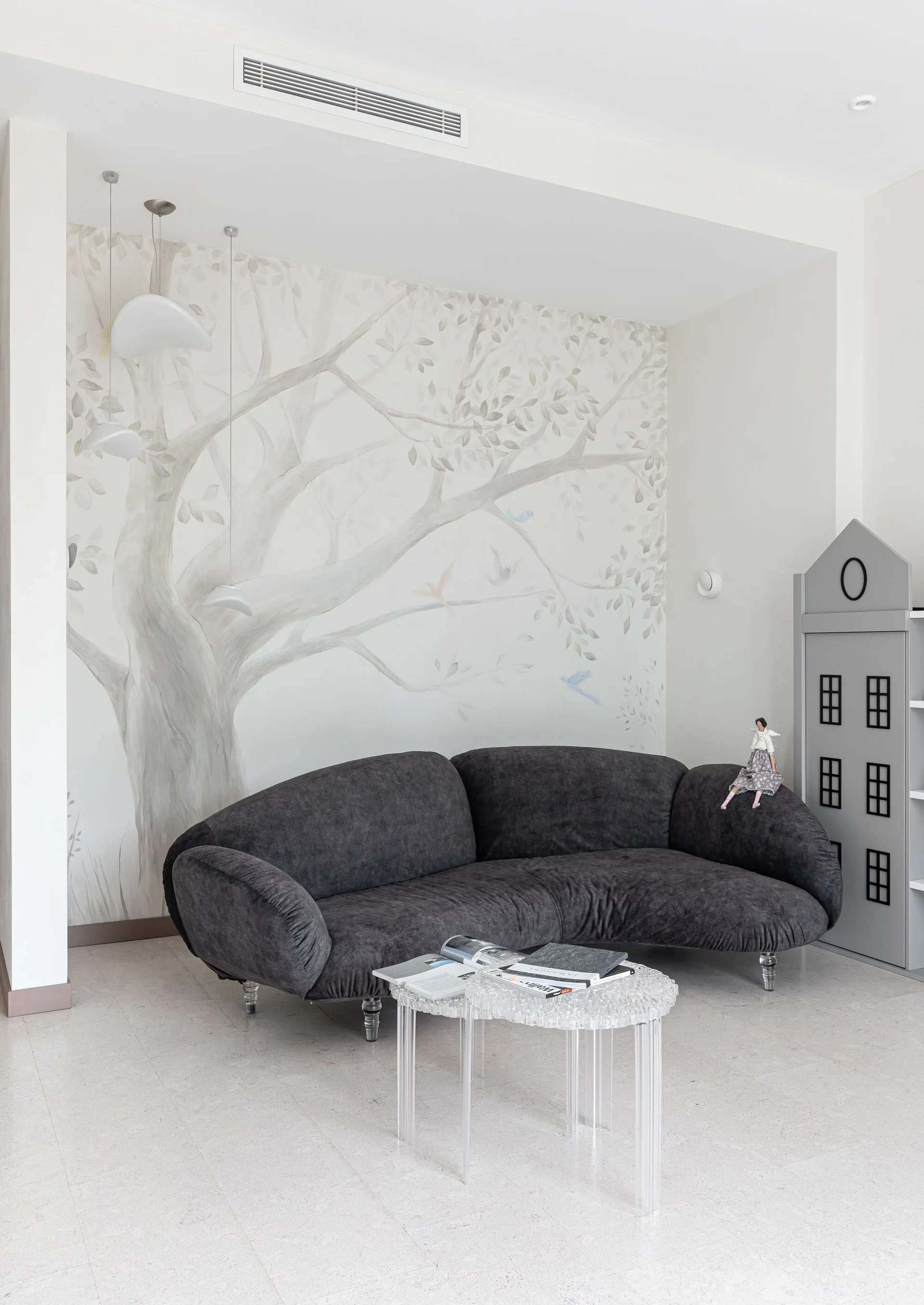 A modern room features a dark gray curved sofa, a clear acrylic coffee table, and a large tree mural on the back wall.