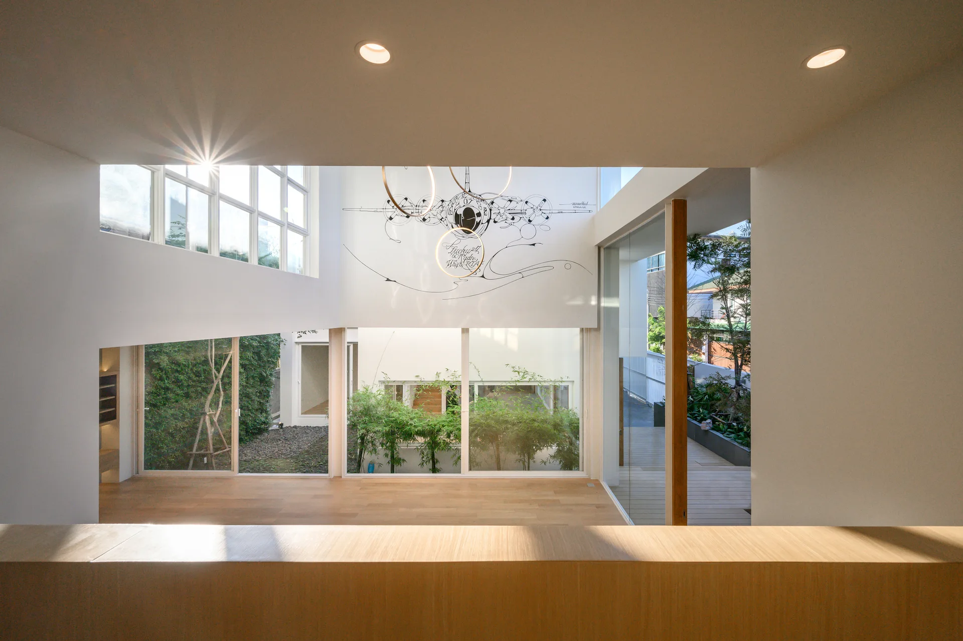 A modern double-height interior with wood flooring, white walls, circular ring pendant lights, and large glass windows overlooking a green courtyard.
