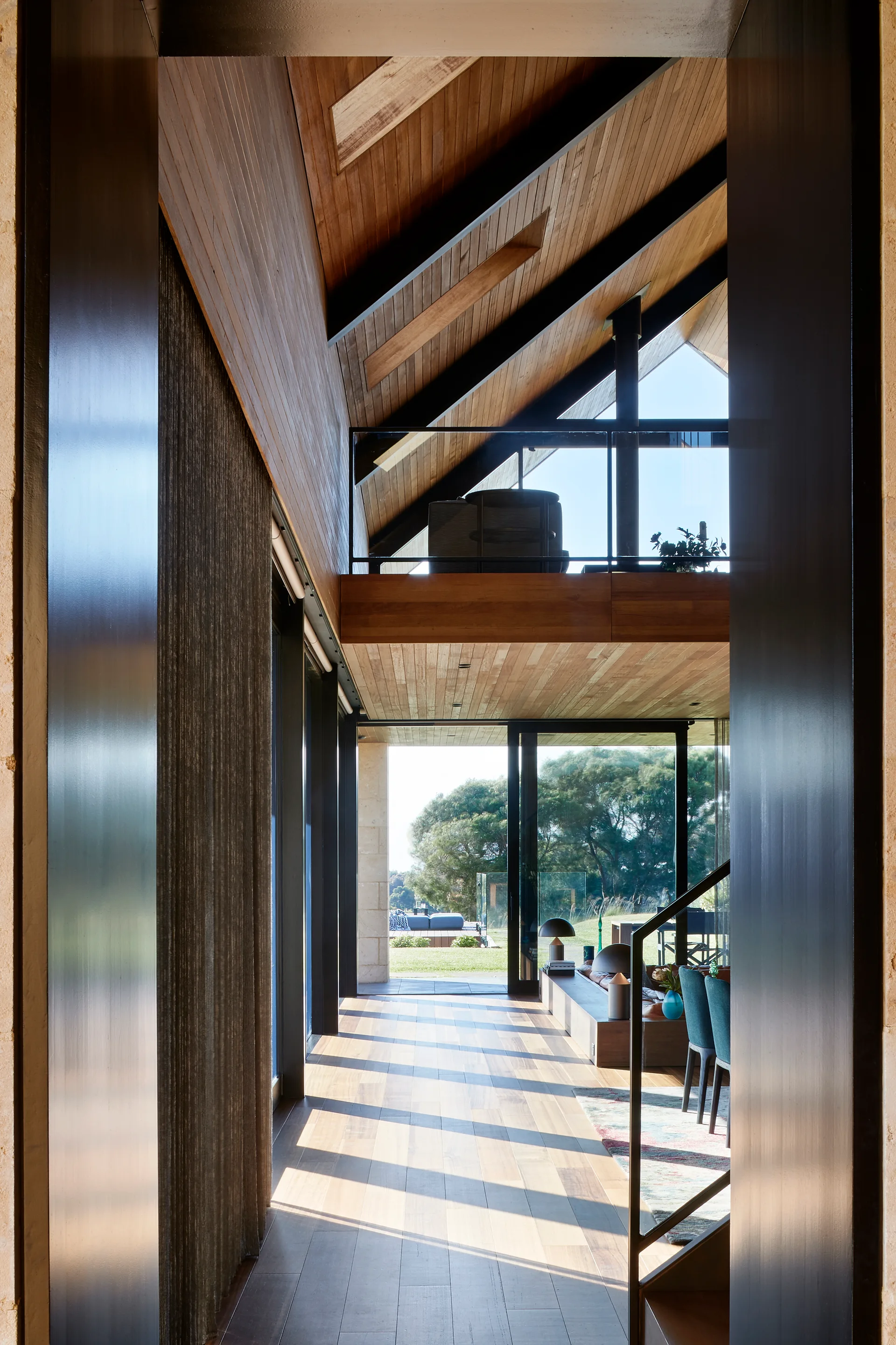 An open-concept living area viewed from a hallway, featuring a sloped wood ceiling with steel beams, a mezzanine loft, and glass doors opening to a yard.