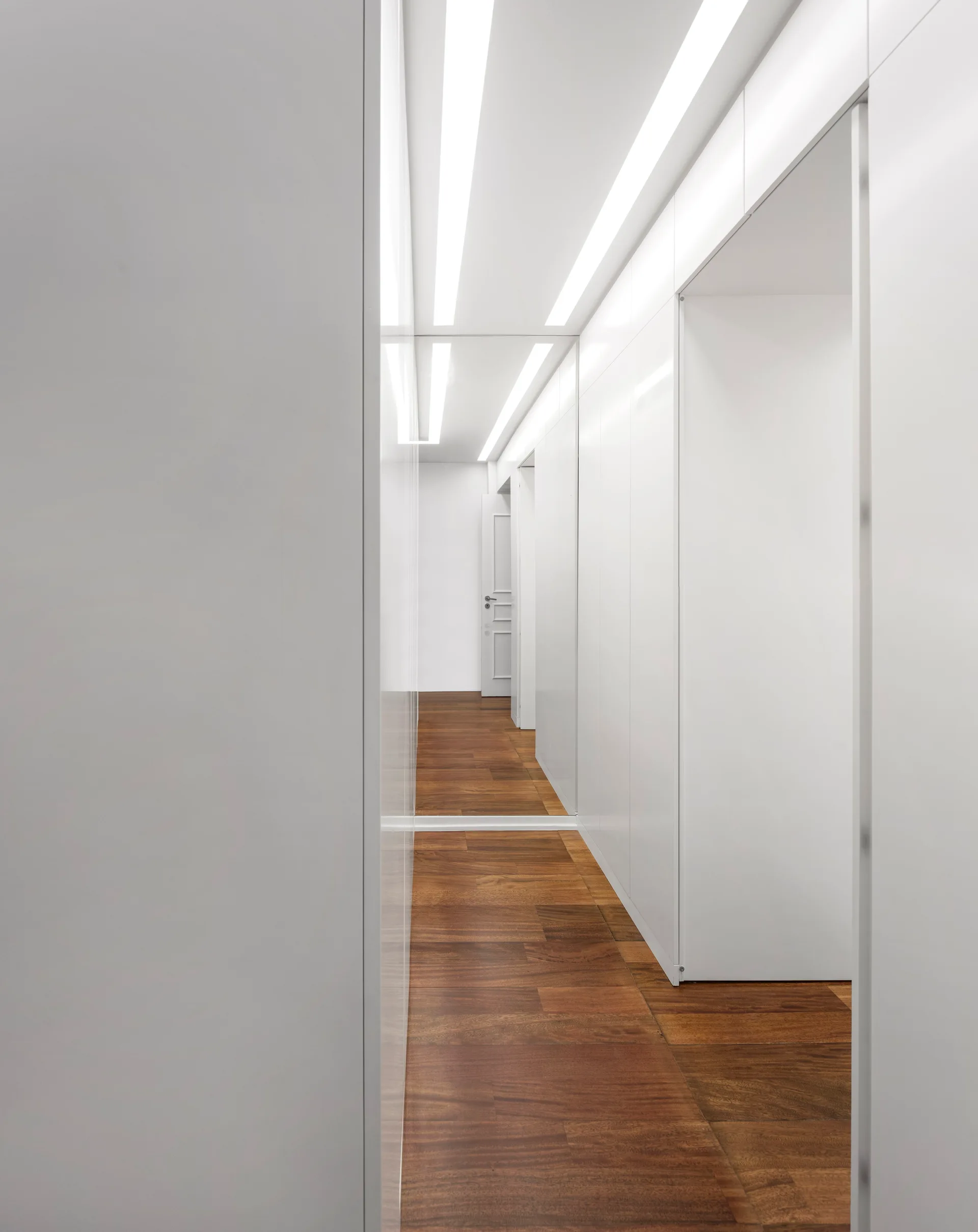 A narrow white hallway featuring floor-to-ceiling glossy cabinetry, recessed linear light strips, and rich brown wood plank flooring.