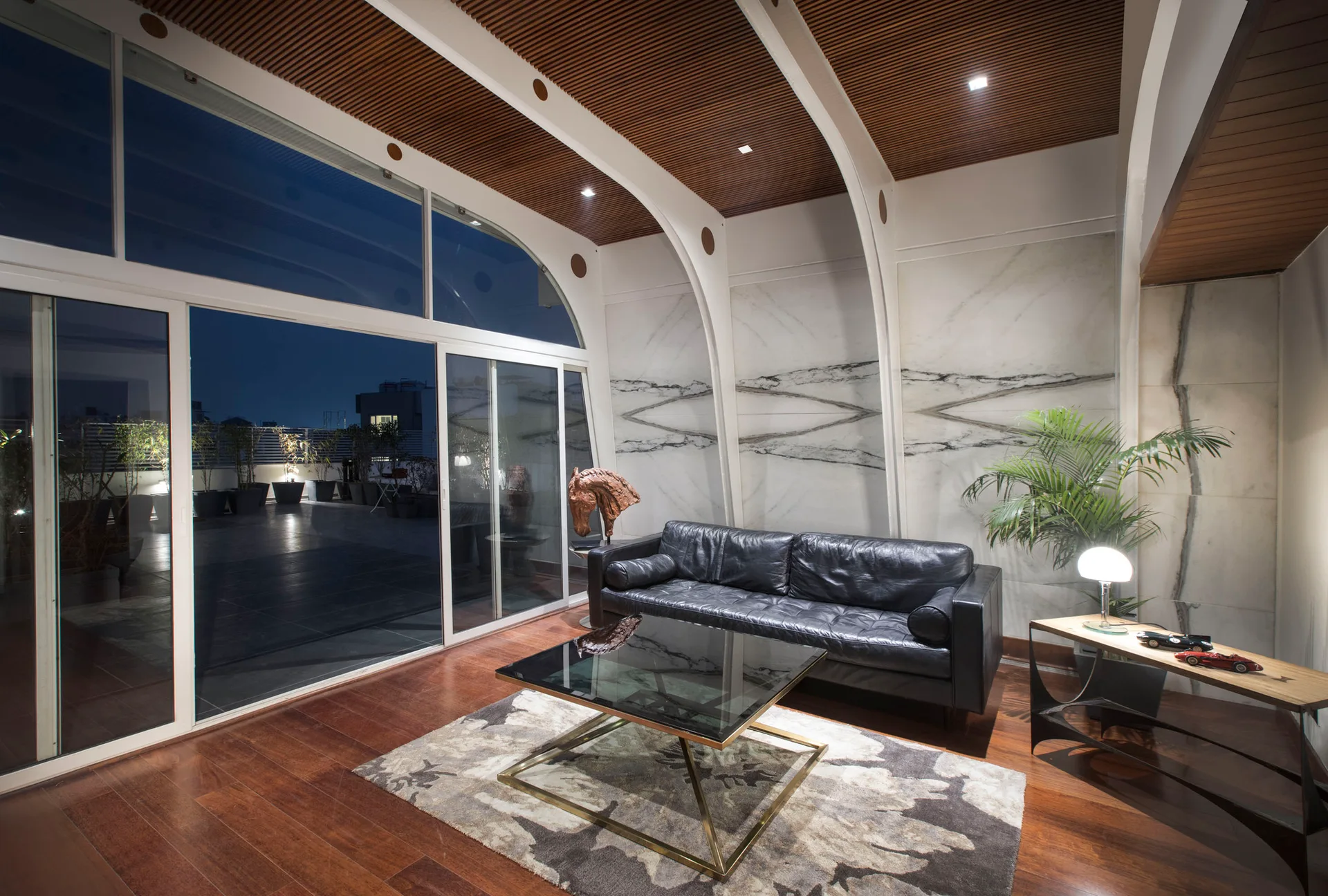 A contemporary living room featuring a black leather sofa, glass coffee table, veined marble walls, and curved white ceiling supports.