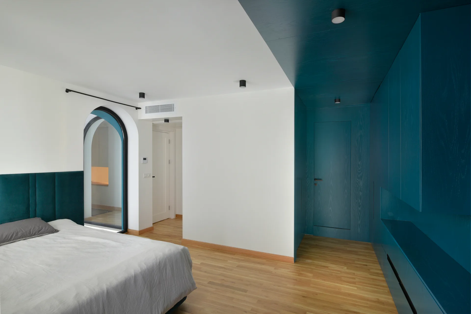 A minimalist bedroom features light wood floors, a bed with a teal headboard, an arched doorway, and deep teal built-in wardrobes.
