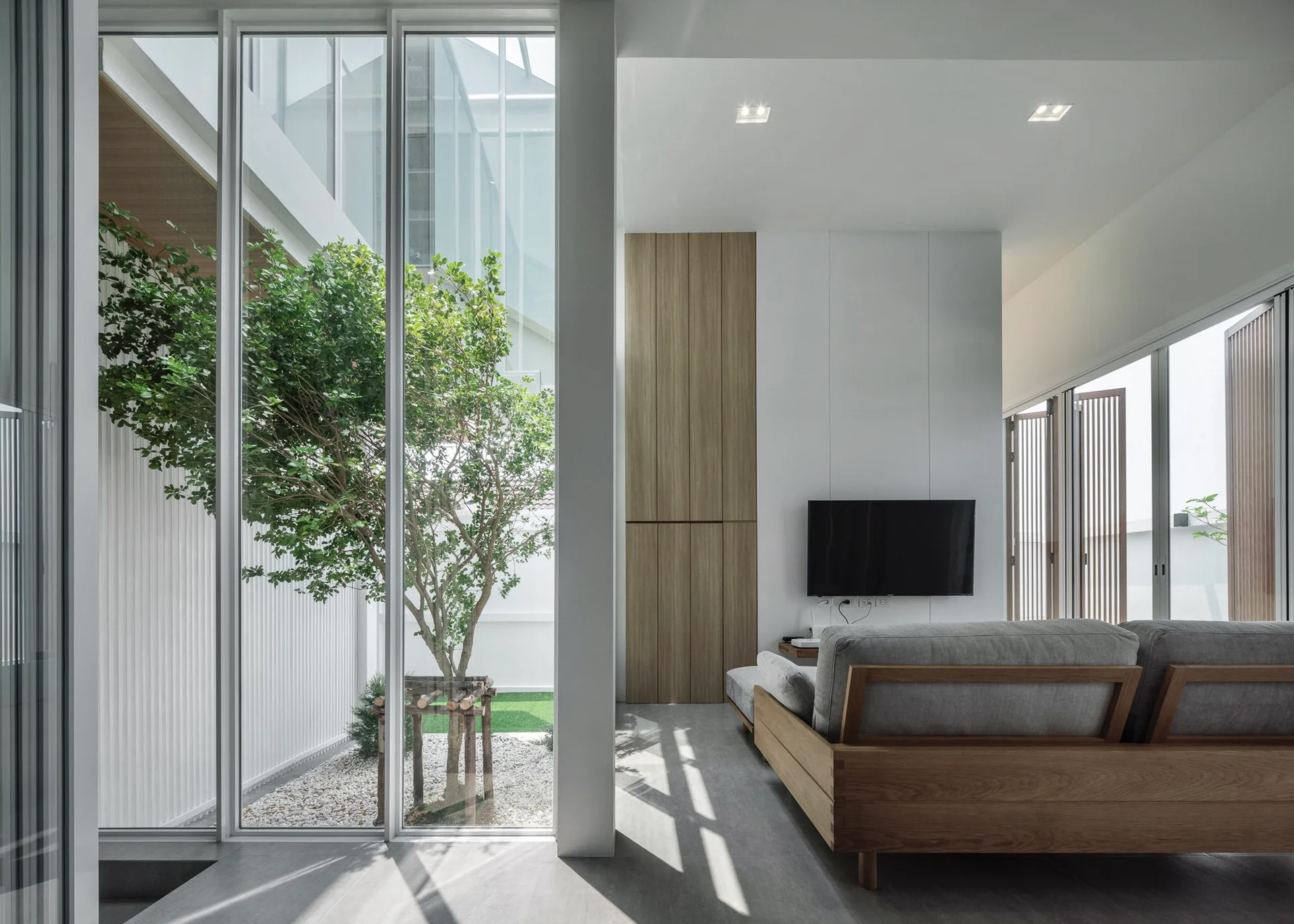 A minimalist living room with a grey sofa and wood cabinetry next to floor-to-ceiling glass walls overlooking an indoor courtyard tree.