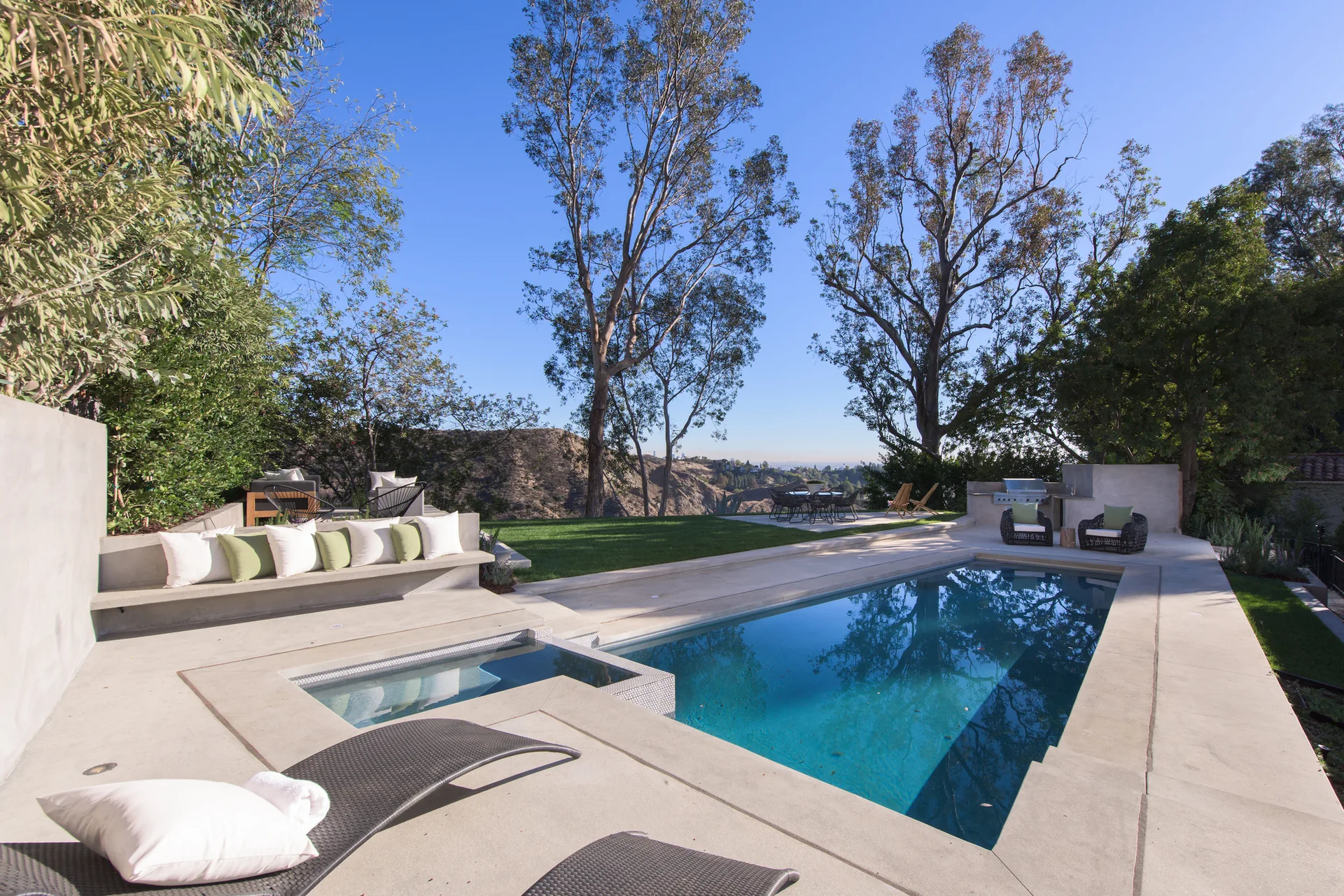 A rectangular swimming pool with an integrated spa is surrounded by a concrete deck, built-in bench seating, and tall trees under a clear blue sky.