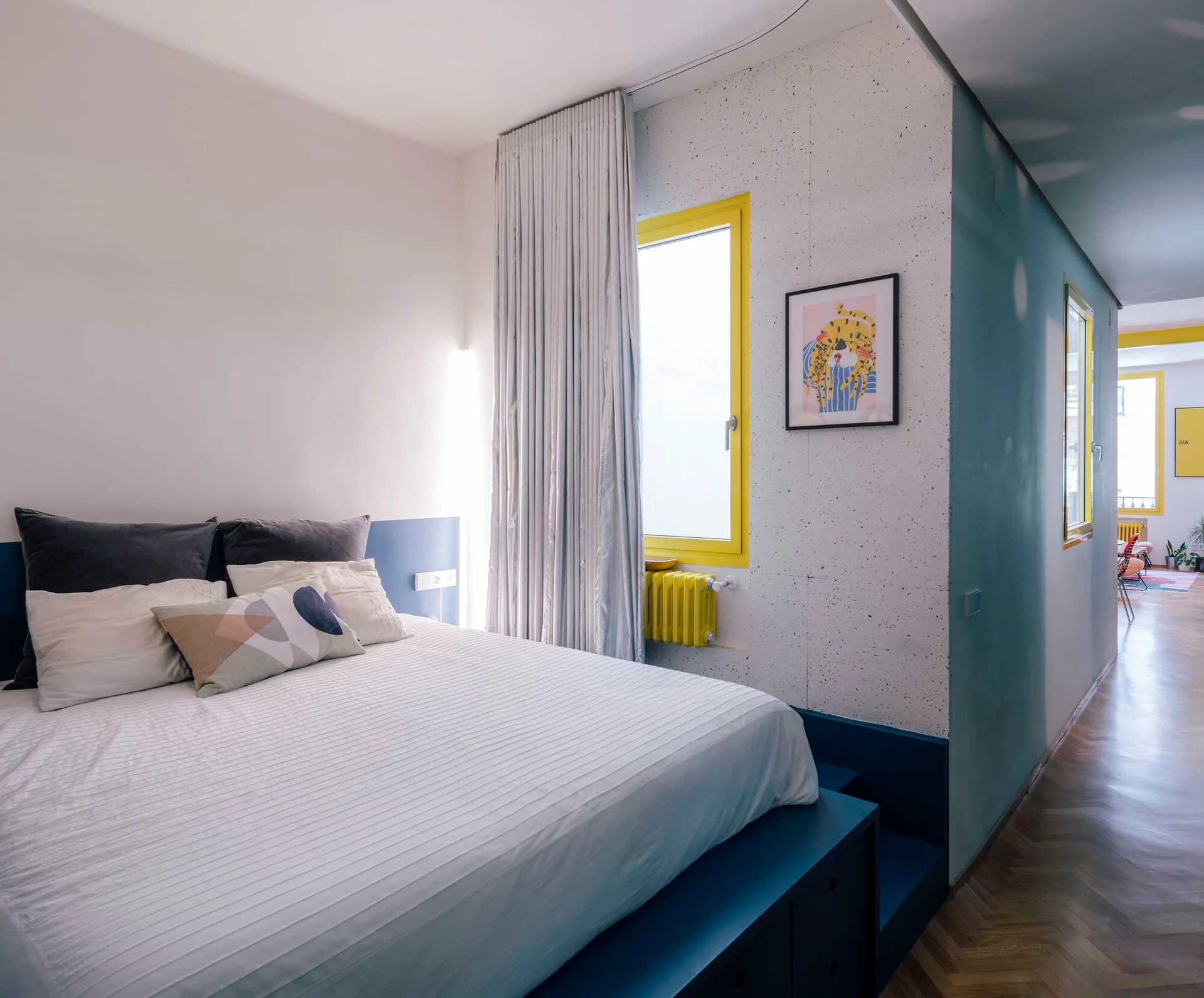 A bedroom featuring a bed on a raised blue platform next to a window with a bright yellow frame and a speckled accent wall.