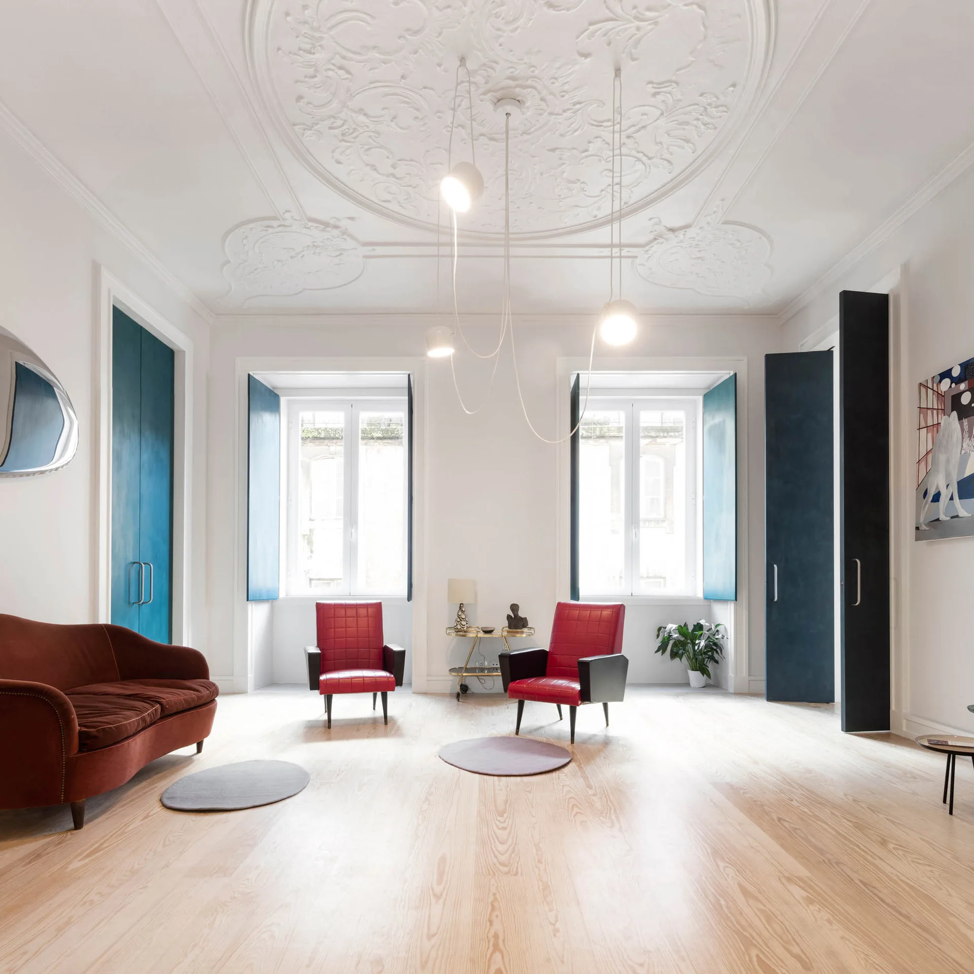 A bright living room with an ornate plaster ceiling, light wood floors, a rust-colored velvet sofa, two red leather armchairs, and tall teal blue doors.