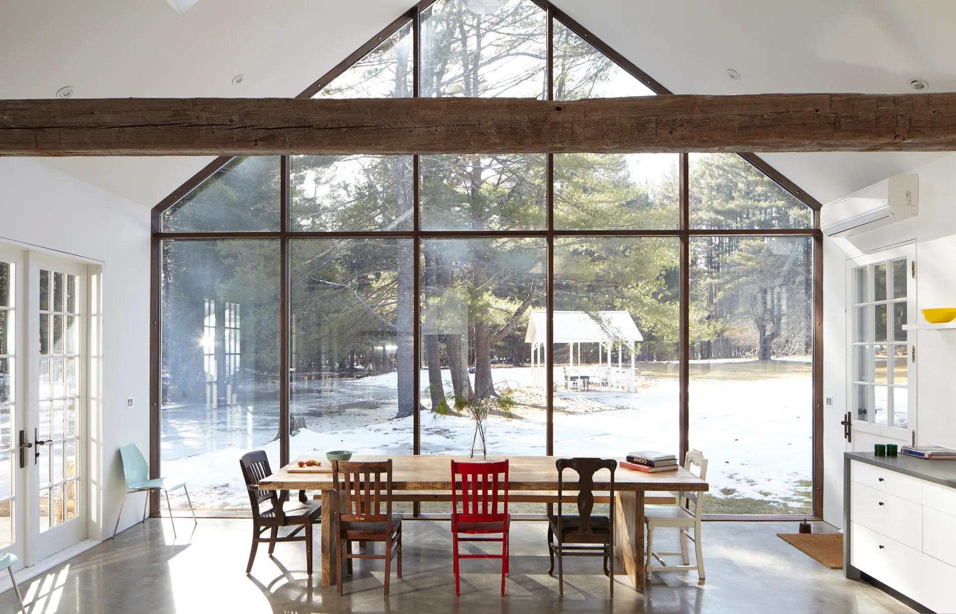 A rustic wooden dining table with mismatched chairs sits on a concrete floor before a massive window wall overlooking a snowy yard.