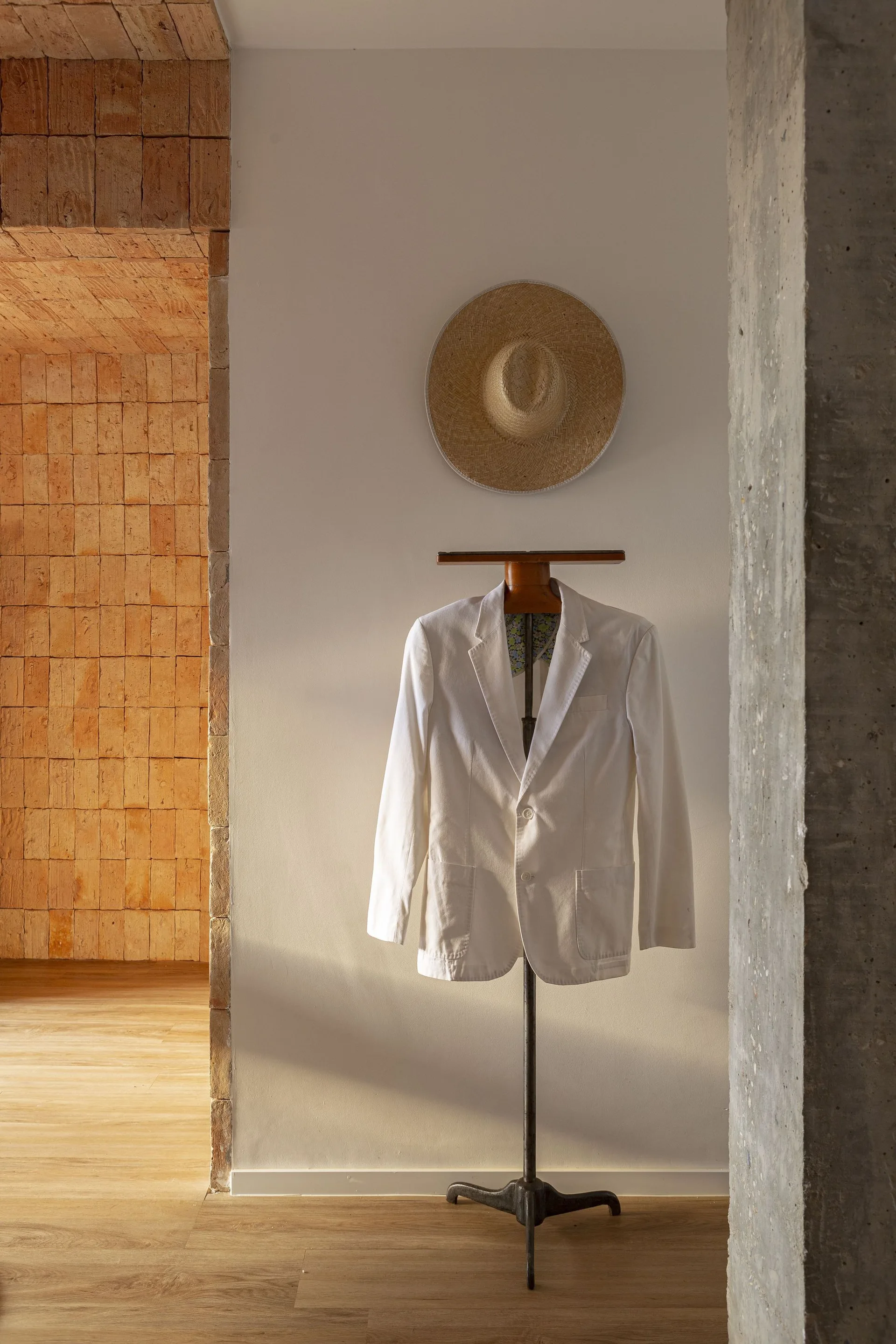 A white jacket hangs on a metal valet stand next to a brick-lined archway and a raw concrete column with a straw hat on the wall.