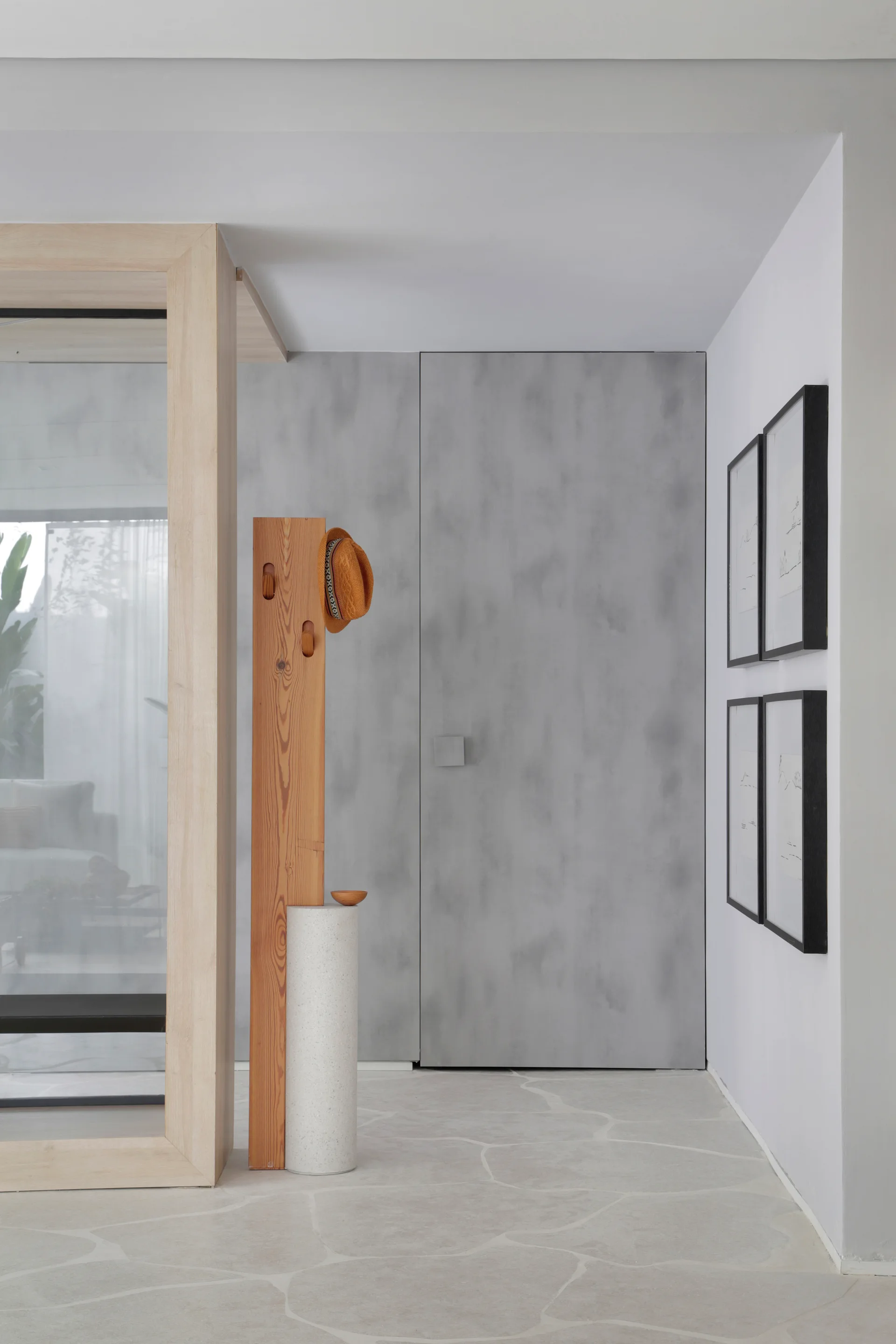 A minimalist entryway features a light wood coat rack on a stone base, a flush grey door, organic stone tile flooring, and framed wall art.
