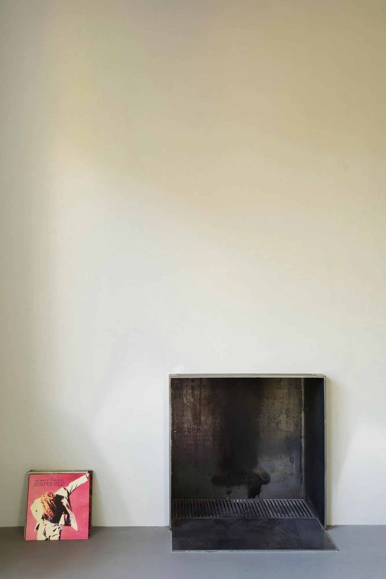 A dark metal fireplace insert sits flush in a smooth off-white wall with a colorful record sleeve leaning on the gray floor.