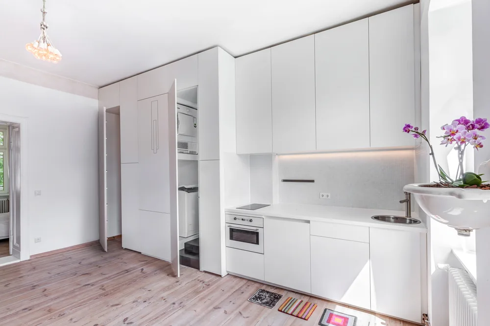 A minimalist white kitchen features handleless cabinets, a small countertop with an oven, and a tall cabinet open to reveal stacked laundry machines.