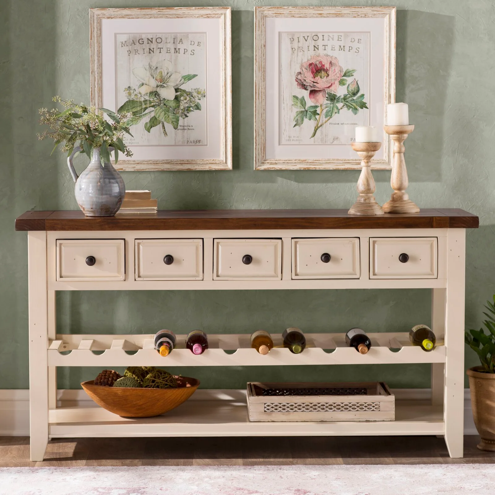 A cream wooden console table with a dark wood top and built-in wine storage is styled against a sage green wall with two botanical prints.