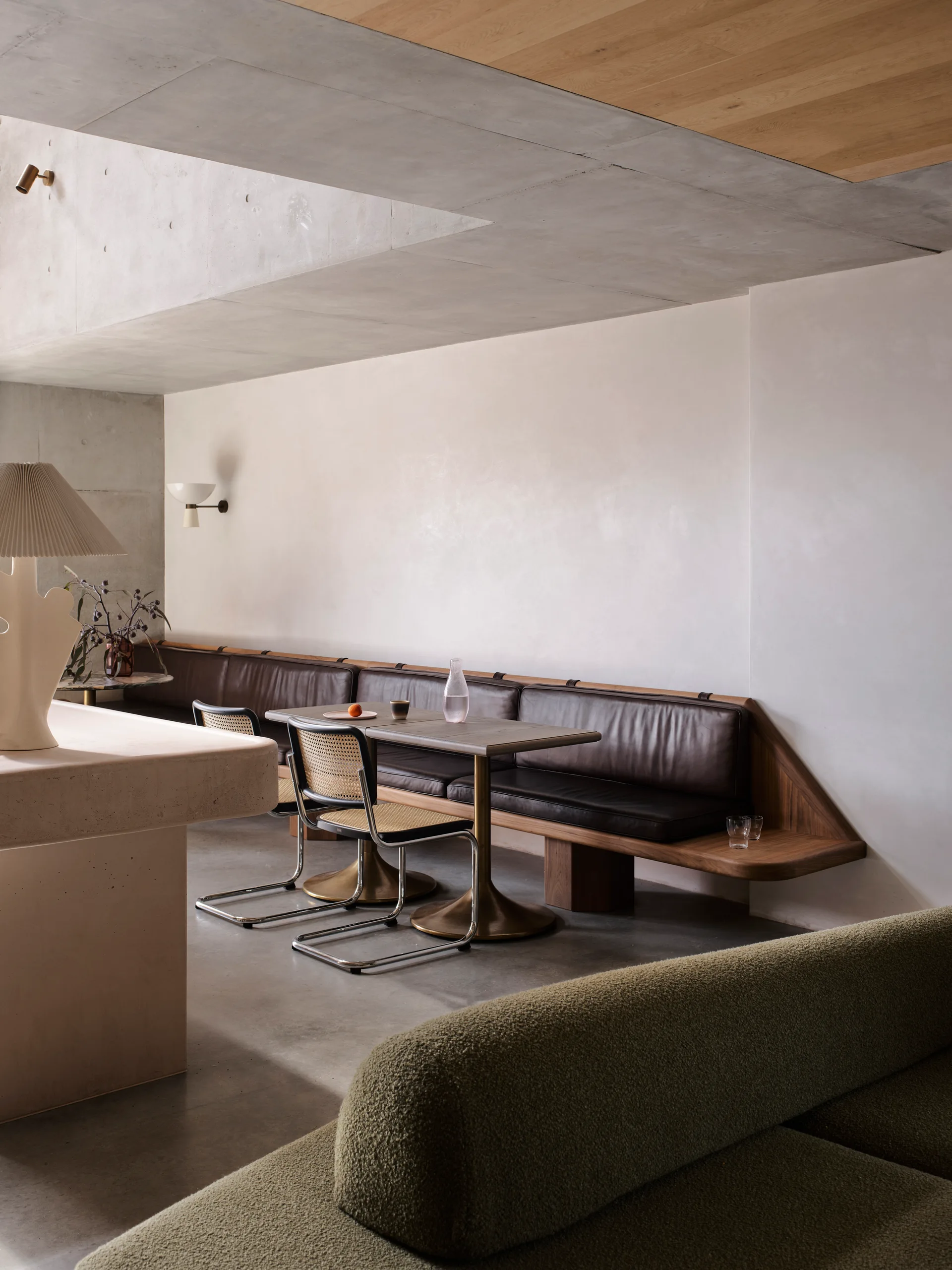 A minimalist dining space featuring a long brown leather banquette, a dark wood table, and cane back chairs beneath a concrete ceiling.
