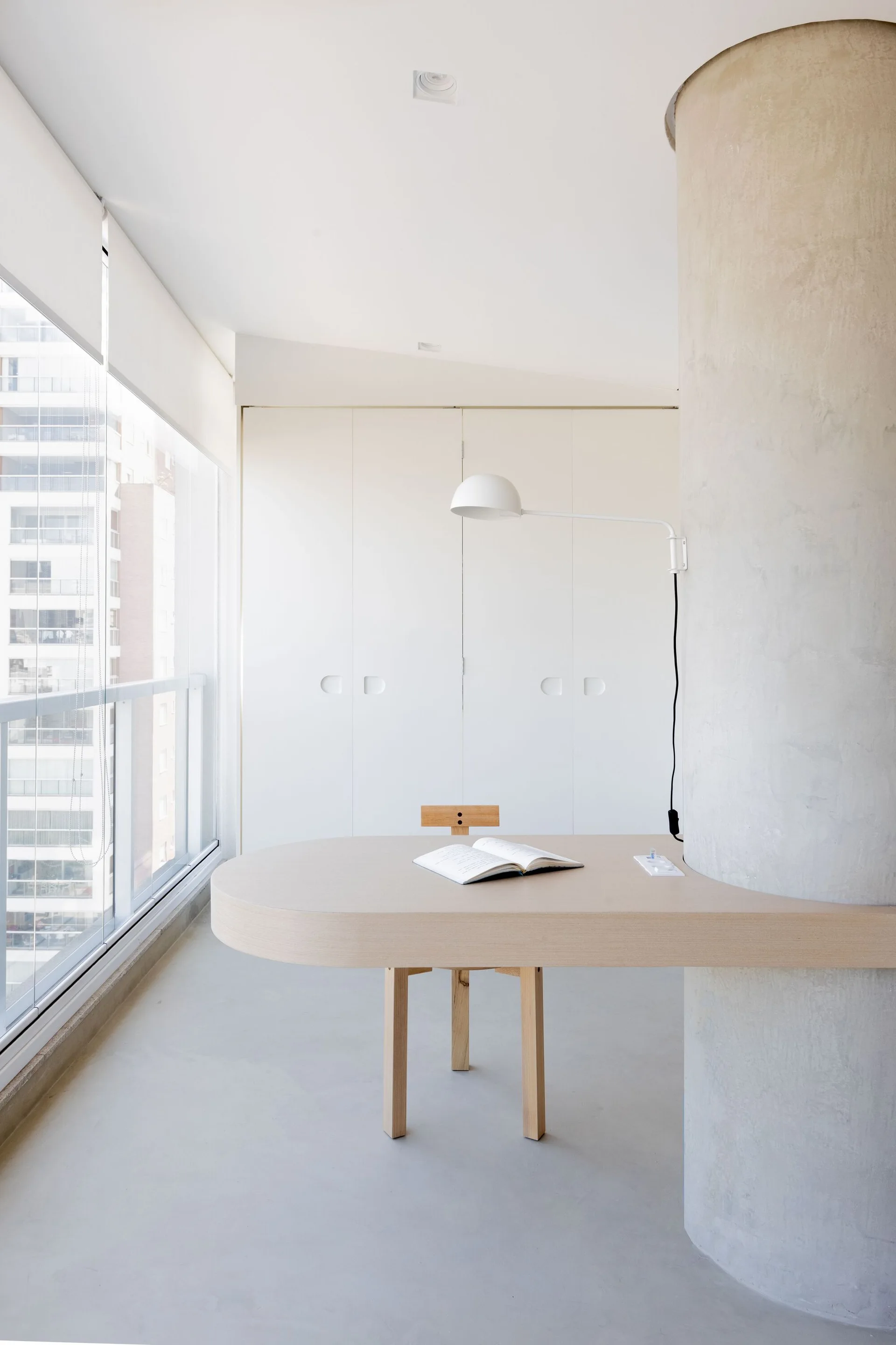A light wood desk wraps around a concrete column in a minimalist home office with white cabinetry and large glass windows.