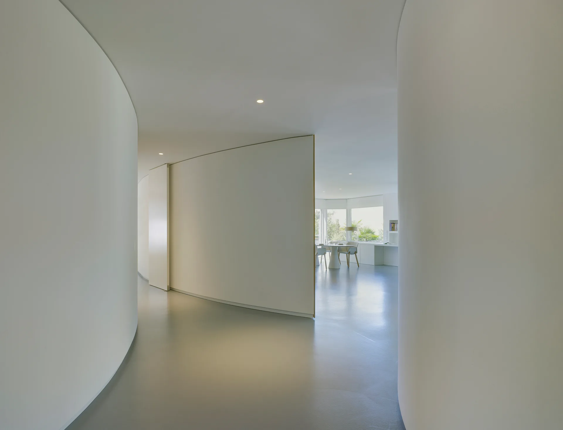 A minimalist hallway features smooth curved white plaster walls and a grey floor leading into a sunlit dining area with a round table.
