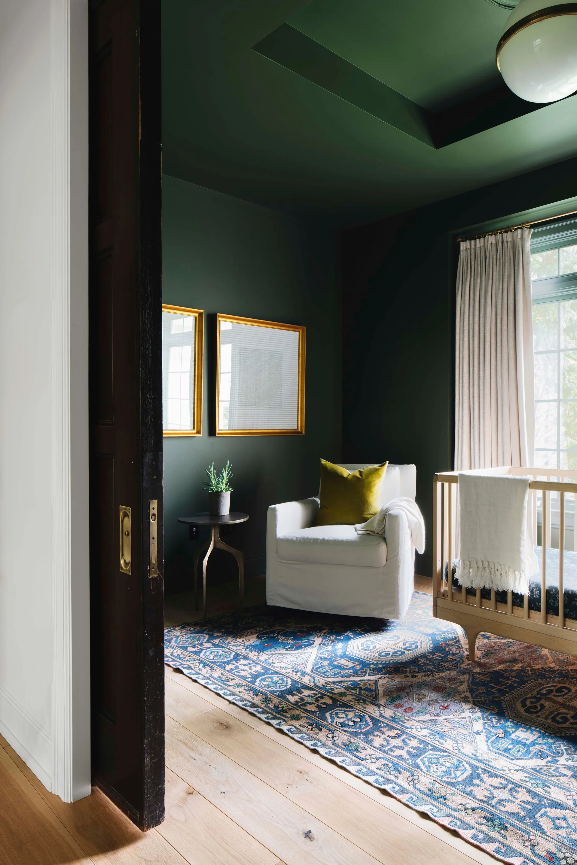 A nursery with dark green walls and ceiling features a wooden crib, white upholstered armchair, and a patterned blue rug.