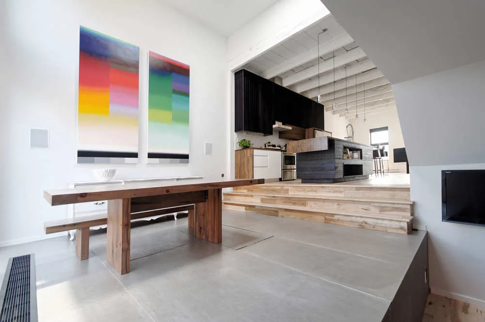 A modern split-level dining area features a large wood table, smooth concrete floors, two tall colorful abstract paintings, and steps leading to a kitchen.