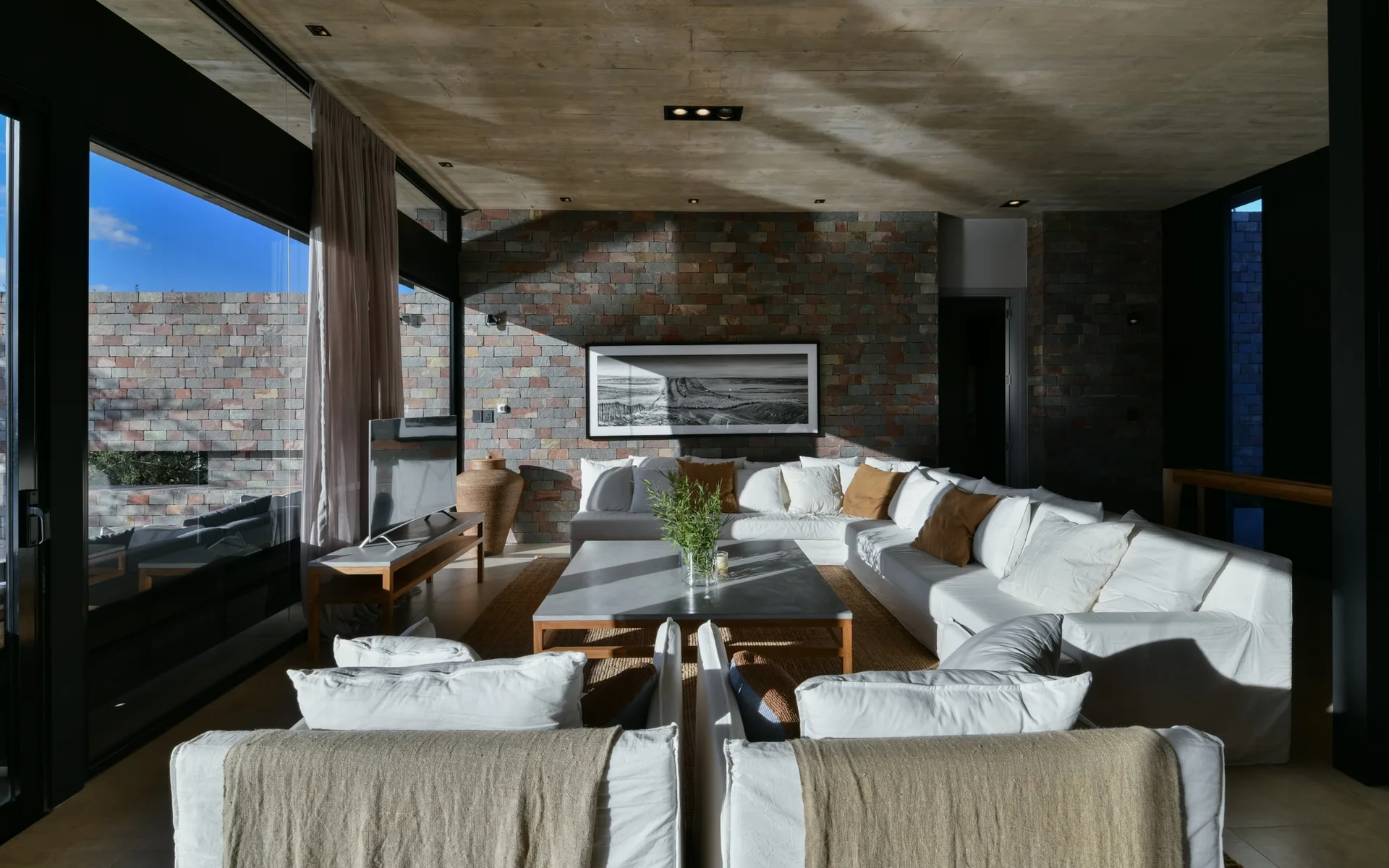 A spacious modern living room featuring a large white sectional sofa, a stacked stone accent wall, a square coffee table, and a raw concrete ceiling.