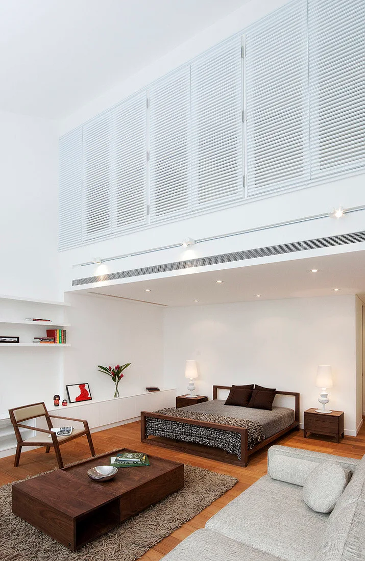 A minimalist loft bedroom features a low platform bed beneath a lower ceiling, with white louvered shutters covering the high upper wall.