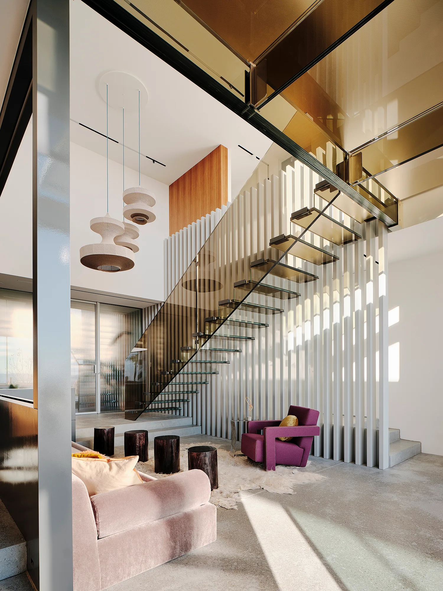 A modern floating glass staircase with a bronze tinted railing is set against a white slatted feature wall next to a pink velvet sofa.