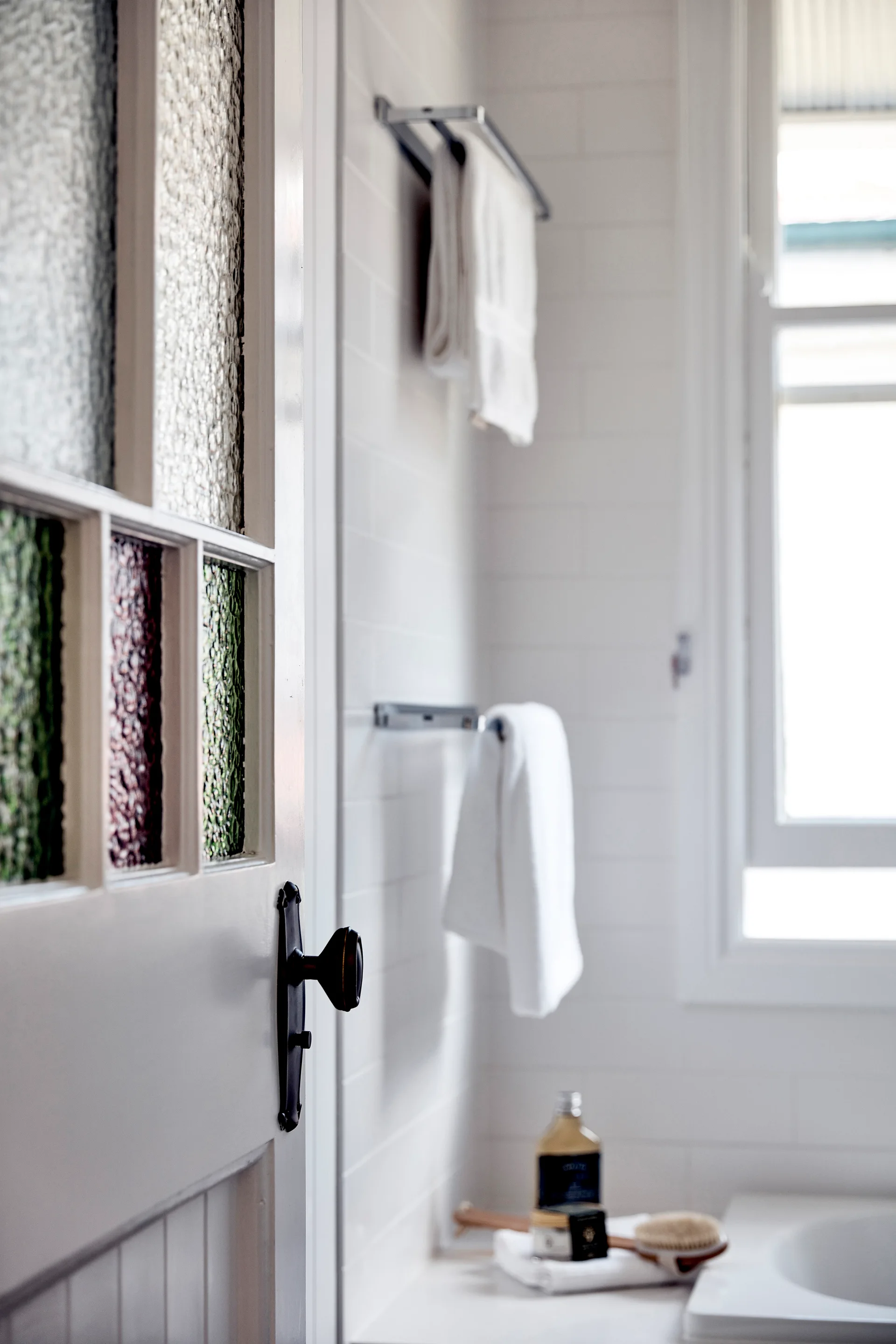 A white door with textured colored glass panes and a black vintage handle opens into a bright white tiled bathroom with hung towels.