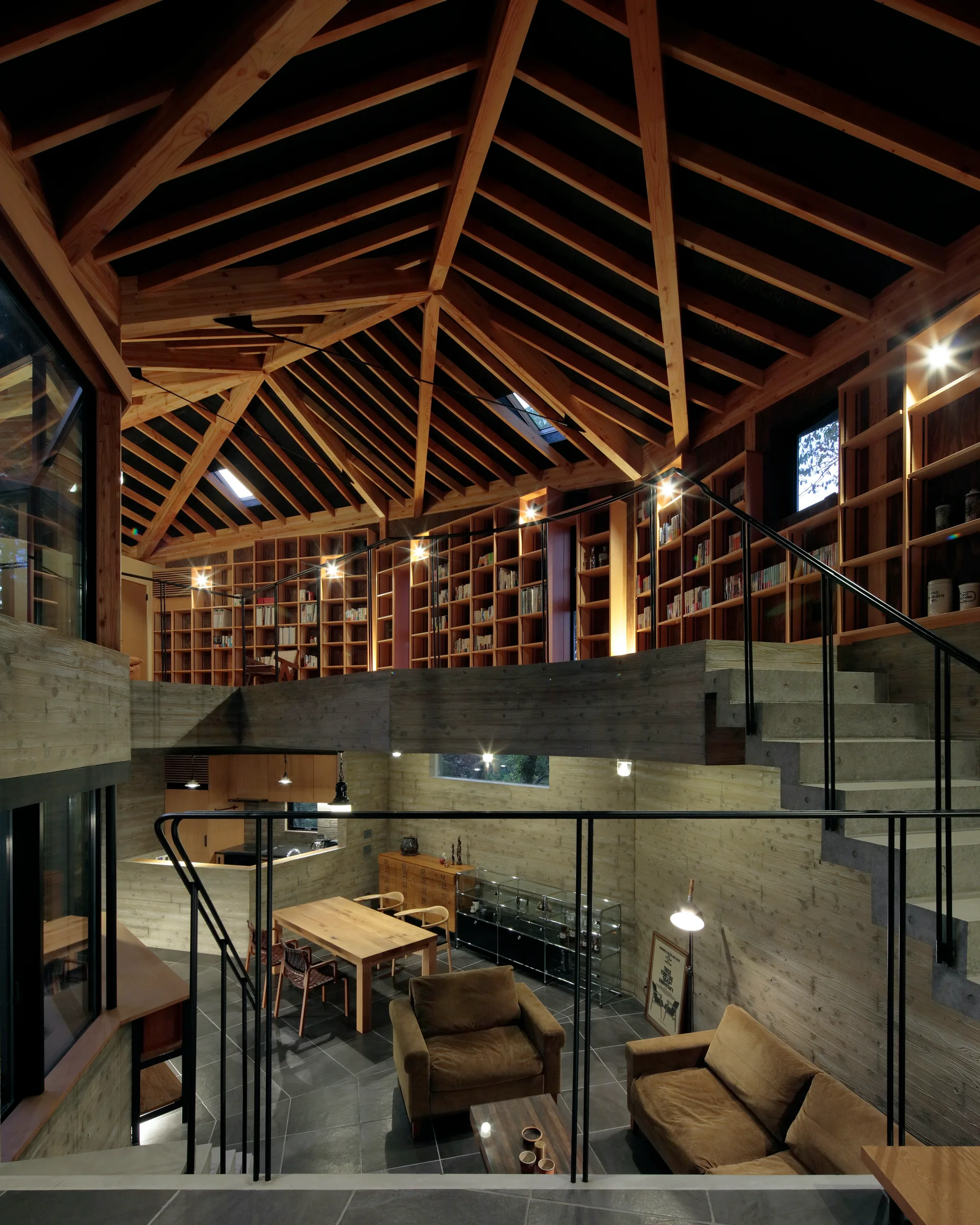 A split-level living area with concrete walls features a dining set, concrete stairs, and a mezzanine library under a geometric timber rafter ceiling.