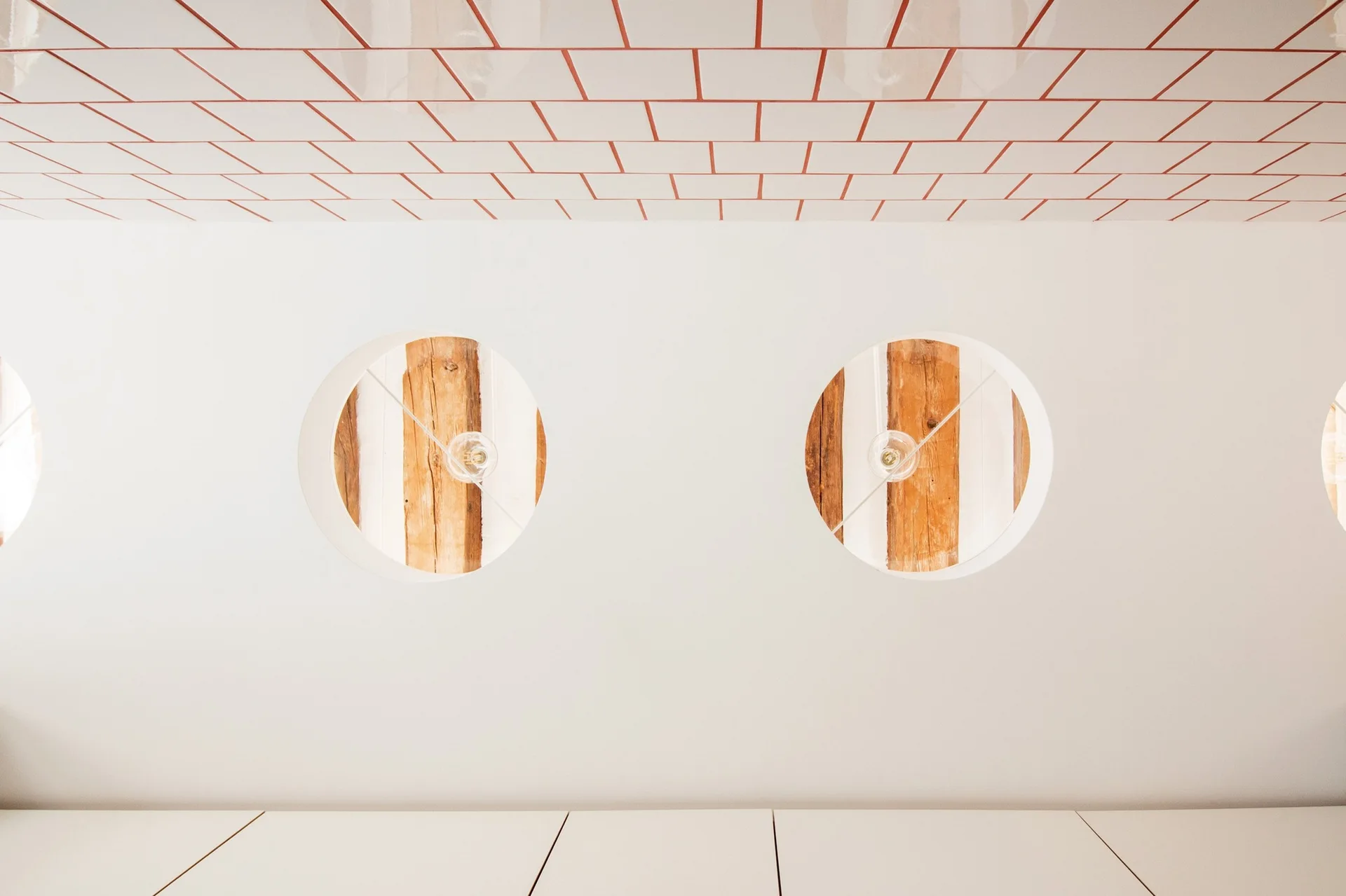 A white wall features two circular cutouts revealing rustic wooden beams and simple hanging lightbulbs beneath a tiled ceiling with orange grout.