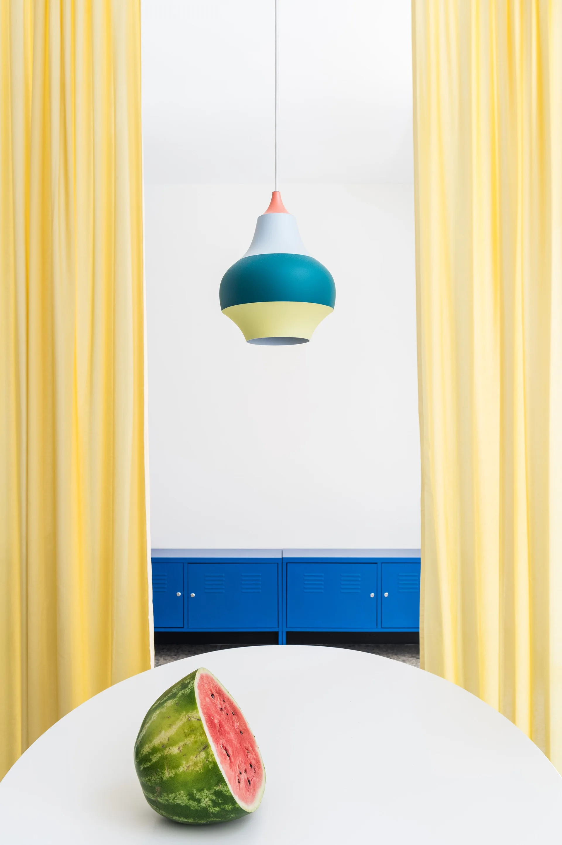 A white table with a watermelon slice is framed by bright yellow curtains, with a blue metal storage cabinet and a hanging colorful pendant lamp.
