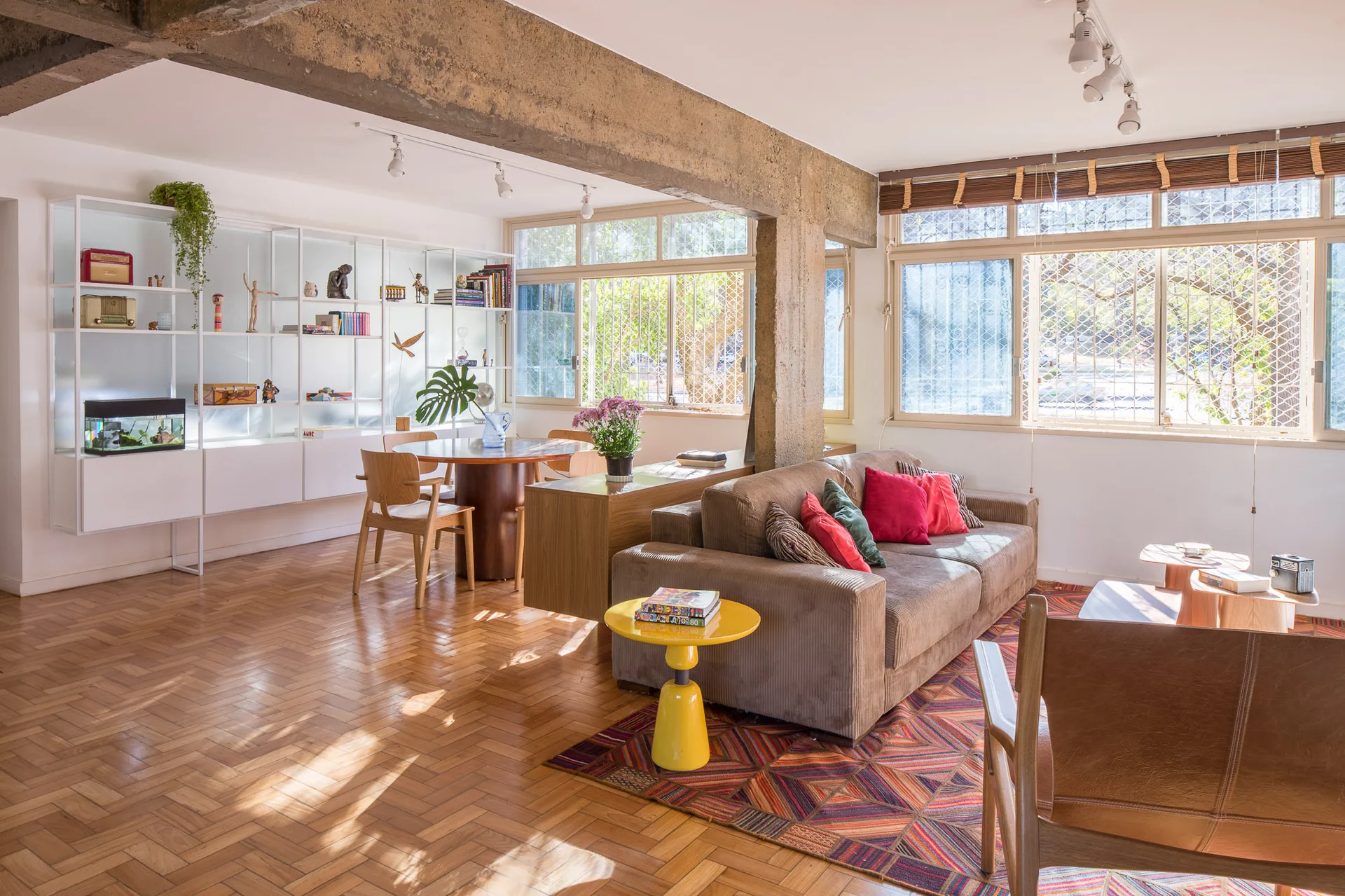 An open-plan living and dining area featuring a brown corduroy sofa, a bright yellow side table, wooden parquet floors, and an exposed concrete beam.