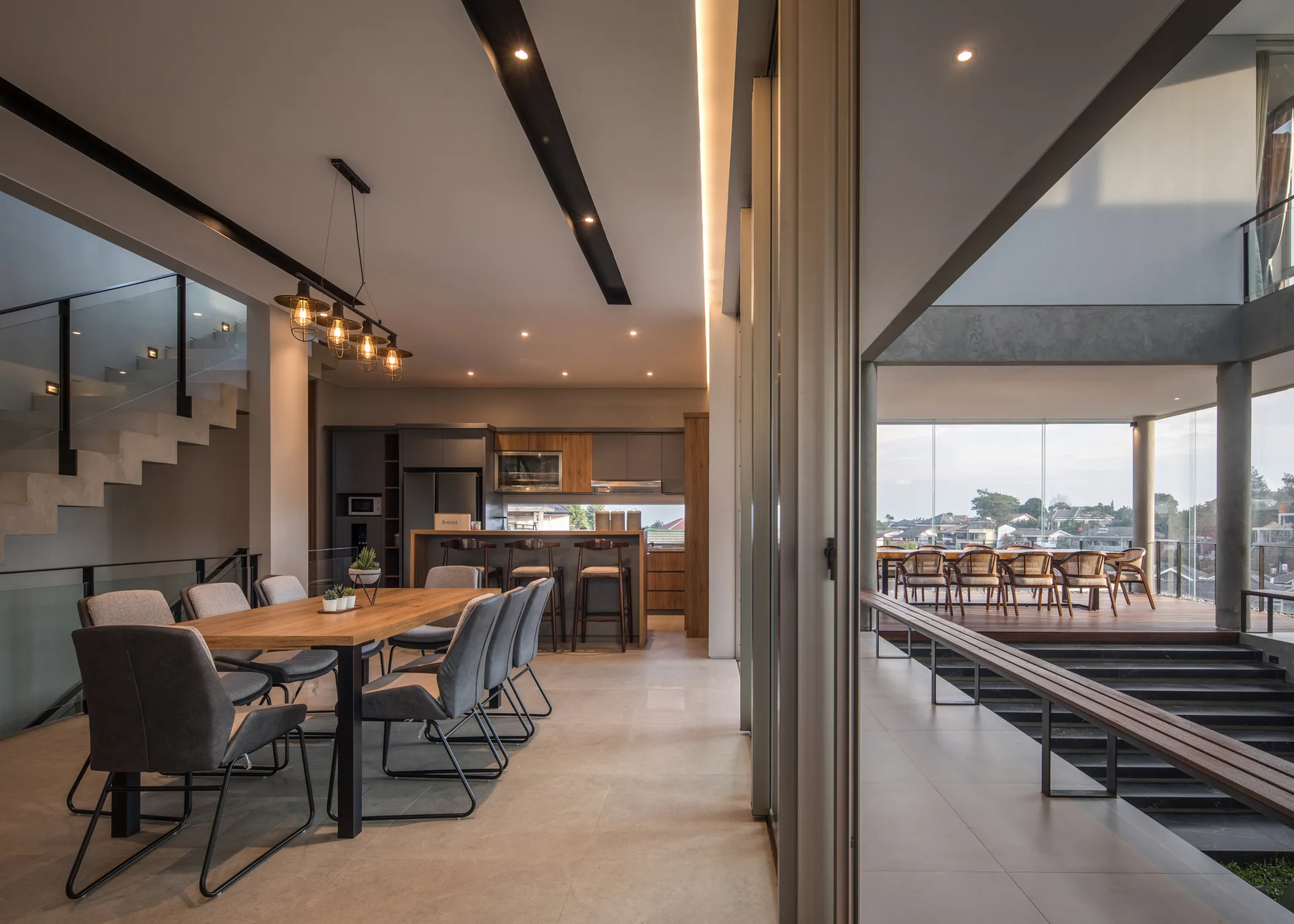 A modern open-plan dining room with a wooden dining table, concrete staircase, and glass walls opening to a wooden terrace.
