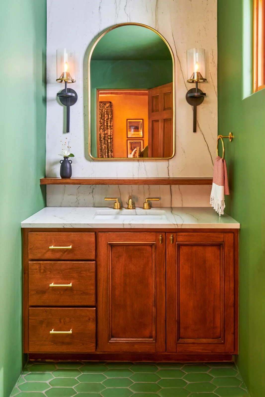 A wooden bathroom vanity with a white marble countertop and backsplash, flanked by green walls and featuring an arched gold mirror and brass fixtures.