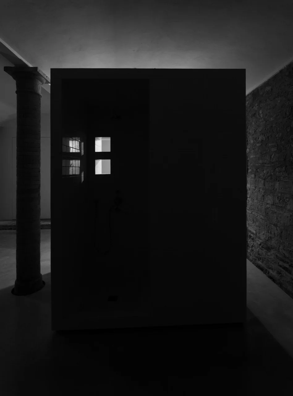 A minimalist dark bathroom featuring a central shower enclosure positioned between a historic stone column and an exposed brick wall.