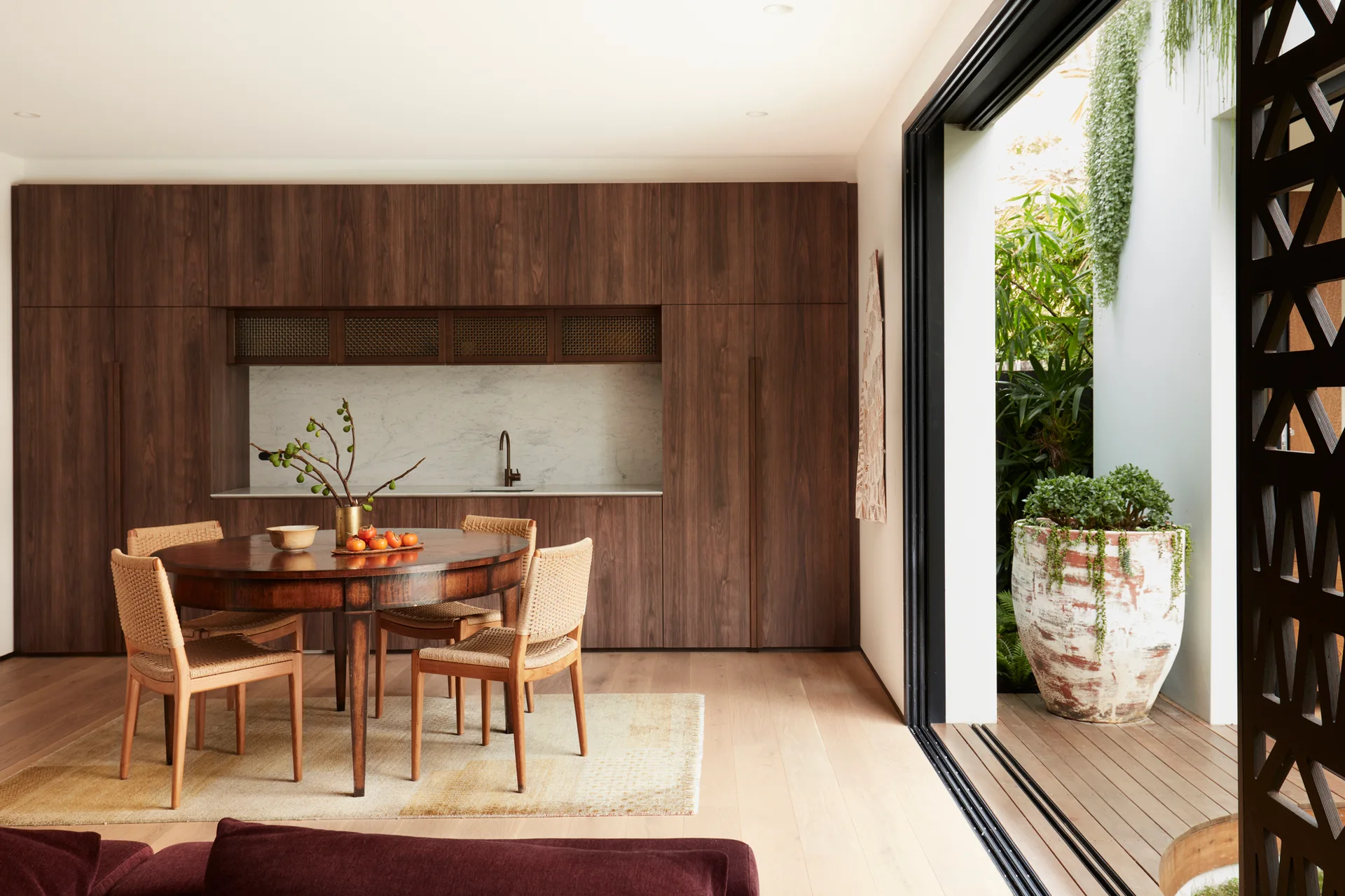 A round wooden dining table with woven chairs sits in front of a walnut-paneled kitchen wall next to a large open sliding glass door.