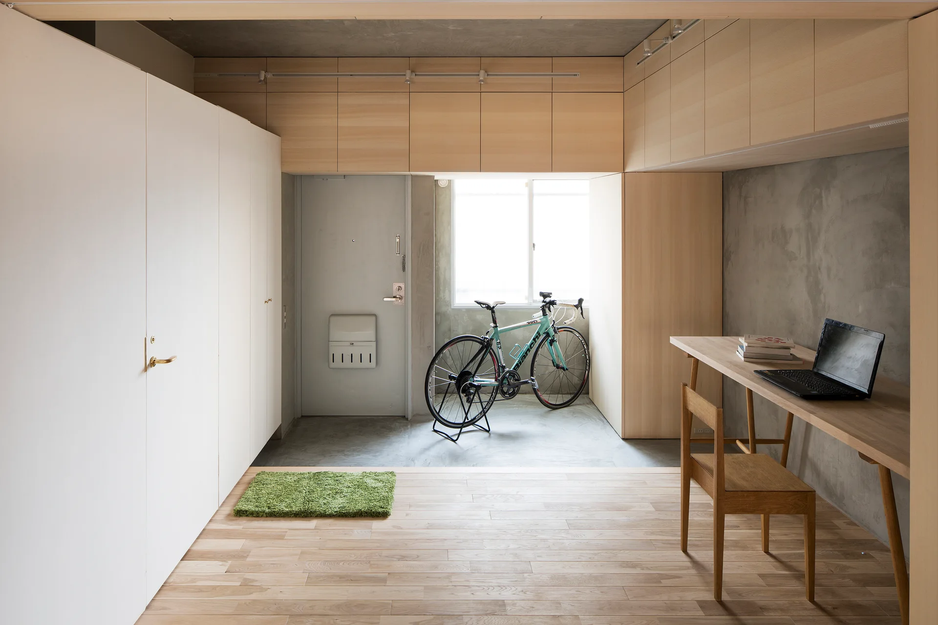 A minimalist entryway with light wood flooring, concrete walls, a wooden desk with a laptop, and a bicycle parked by the window.