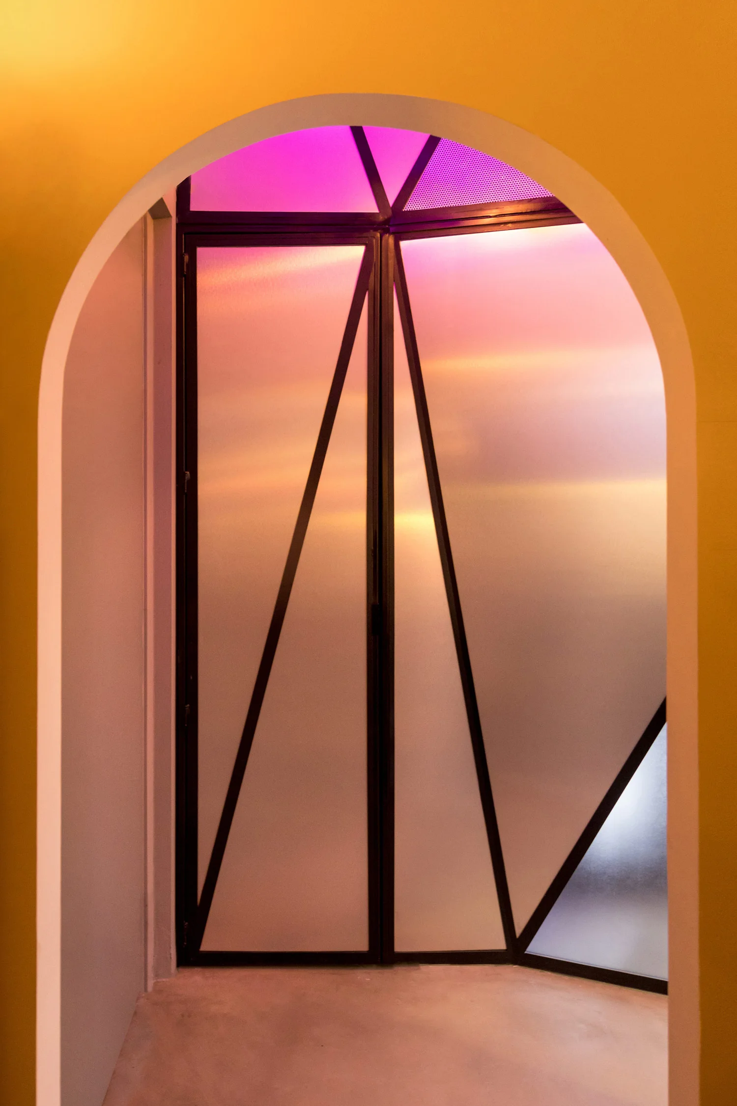 An arched doorway framed in yellow paint reveals a geometric black metal door with frosted glass panels backlit by pink and gold light.