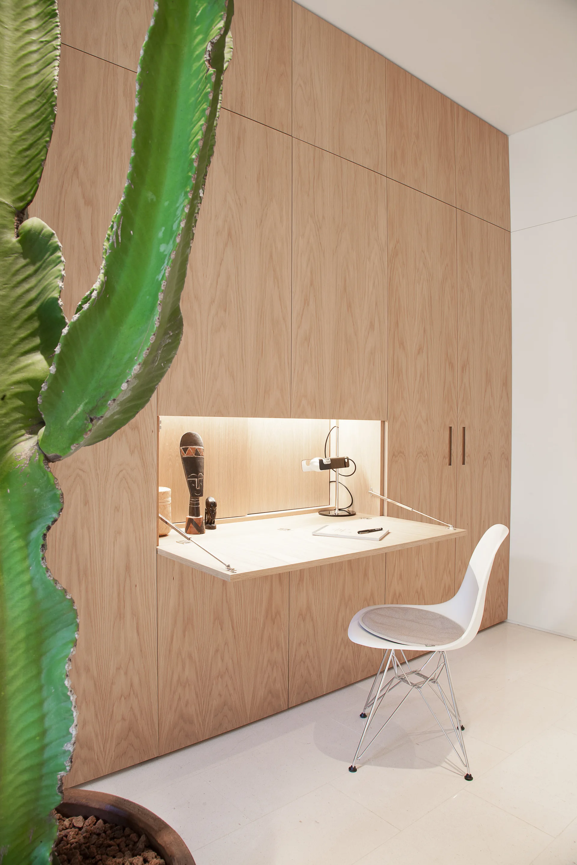 A minimalist wood cabinetry wall features a built-in fold-down desk with warm task lighting, a white chair, and a large green cactus.