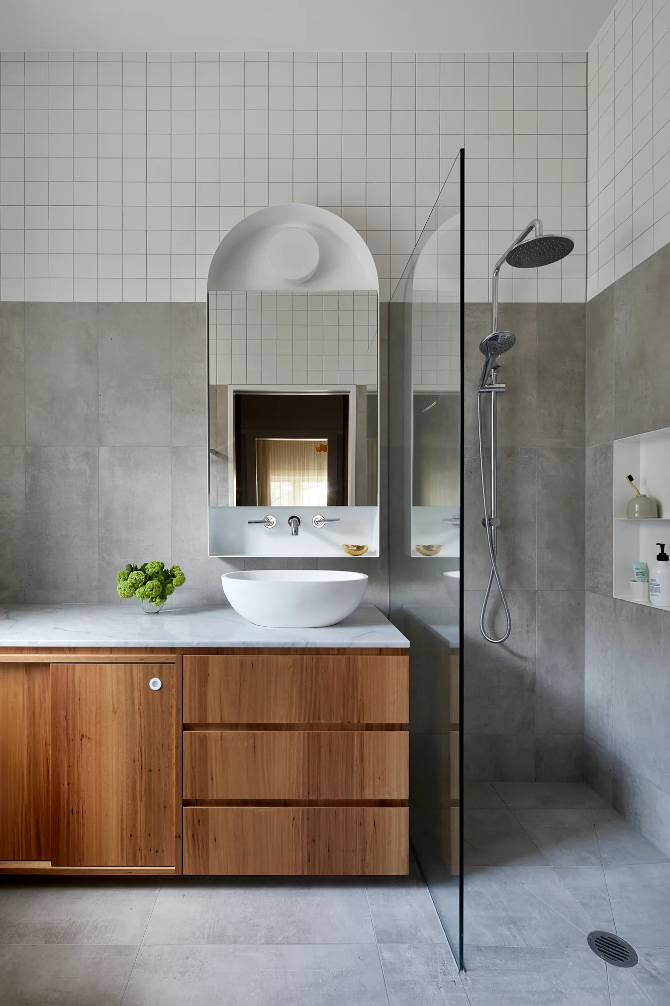A modern bathroom with a natural wood vanity, white vessel sink, arched mirror, and a walk-in shower with grey and white tiling.