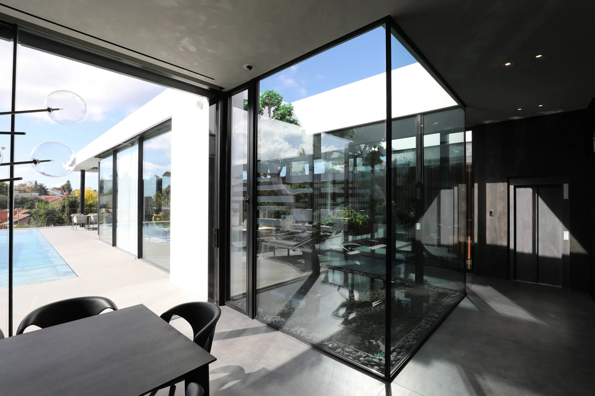 A modern dining room with a black table features floor-to-ceiling glass walls, a glass-encased indoor courtyard, and an outdoor swimming pool.