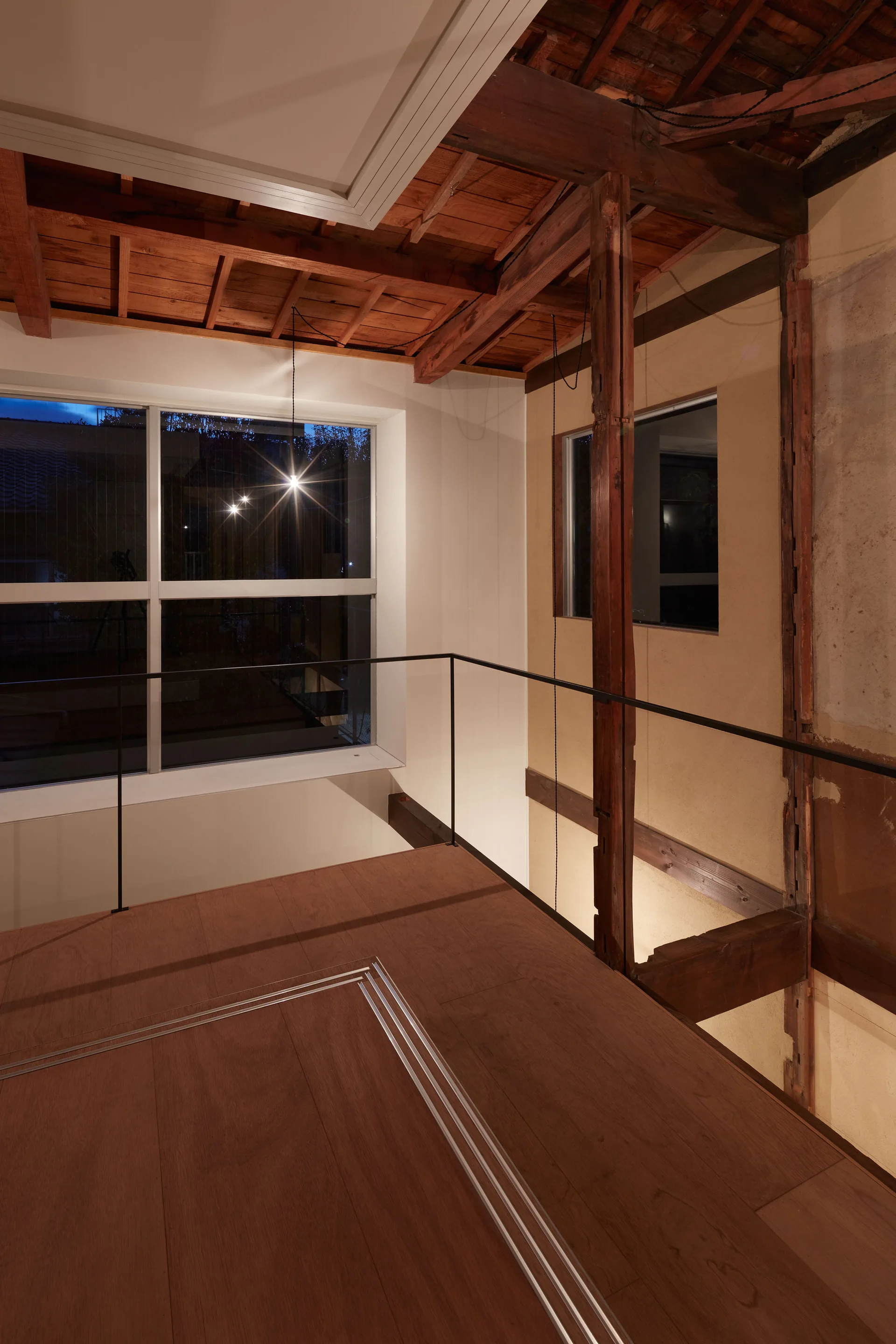 A modern wooden mezzanine landing features an ultra-slim black metal railing, exposed rustic ceiling beams, and a large window at dusk.